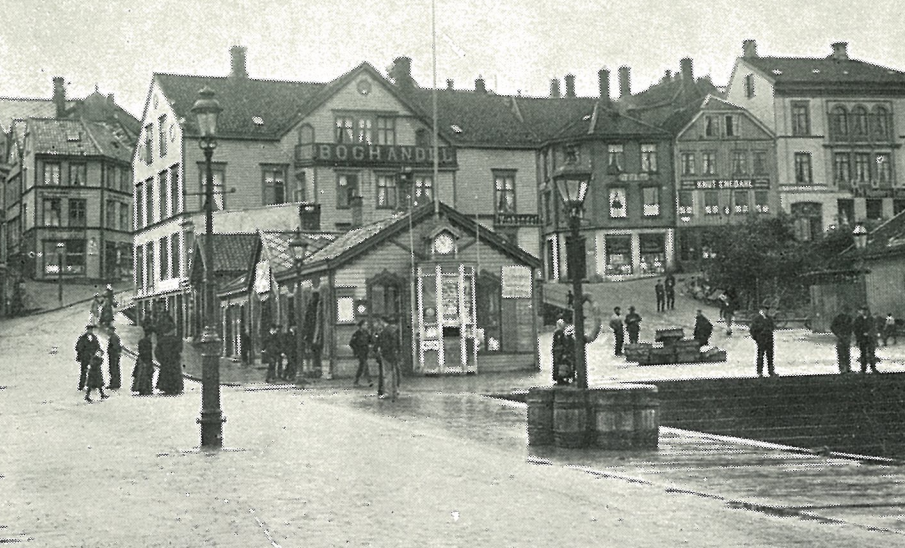 Historical black-and-white photo of a busy city square with a small ticket booth at the center, surrounded by people, street lamps, and multi-story buildings in the background.