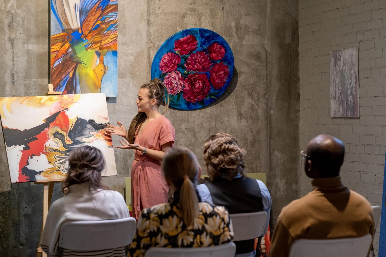 An art instructor explaining a painting to an audience in an art gallery. The viewer's perspective includes three seated people listening. The gallery has concrete and brick walls decorated with colorful paintings, including an abstract piece and a floral artwork.