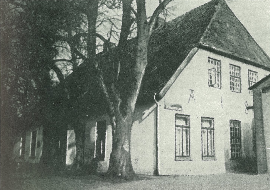 A black and white photo of a large house with a steep gable roof and multiple small windows, with a large tree in front.
