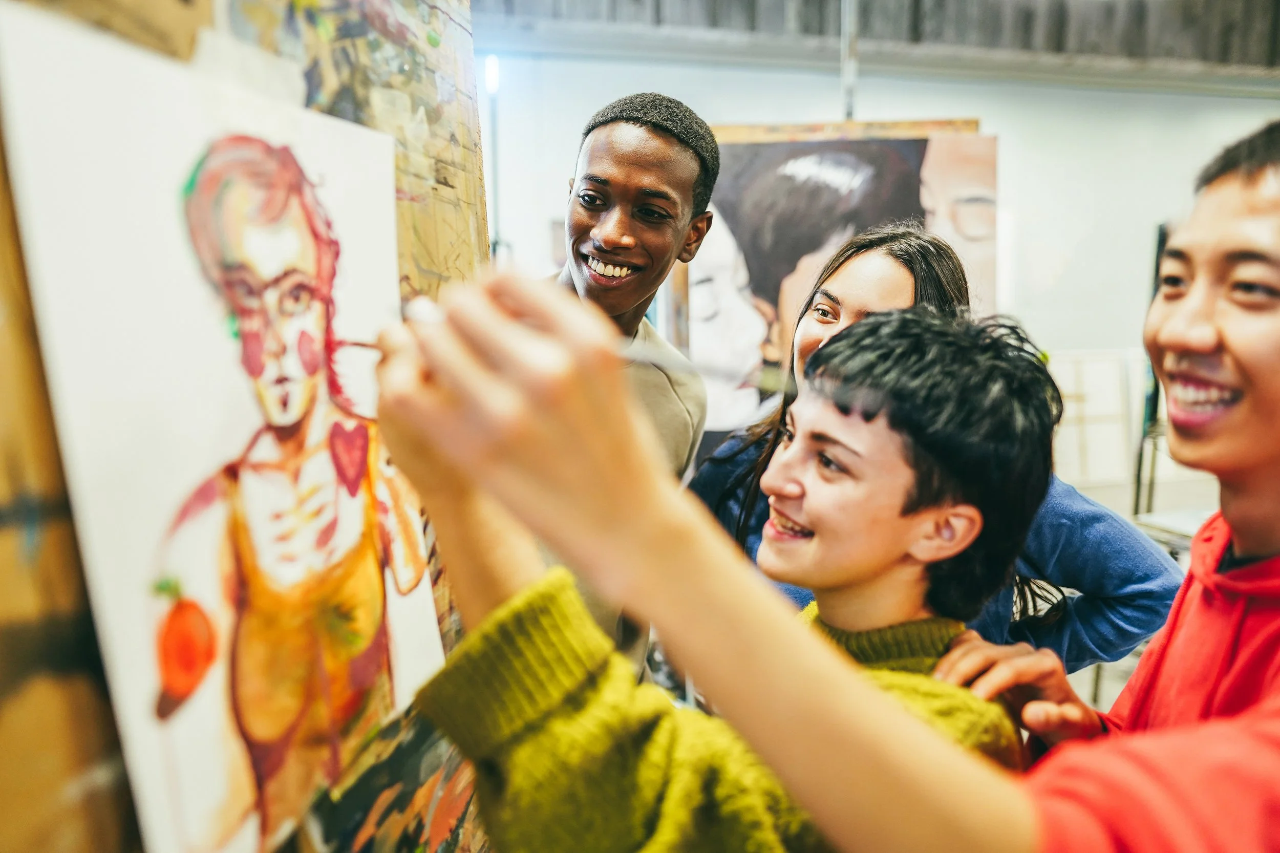 A group of young people gathered around an art canvas, smiling and observing as one person paints a colorful portrait.