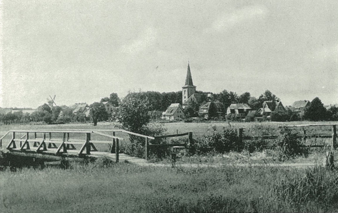 A black-and-white photo of a small rural village with a church with a tall steeple, surrounded by houses, a windmill in the background, and a wooden bridge in the foreground.