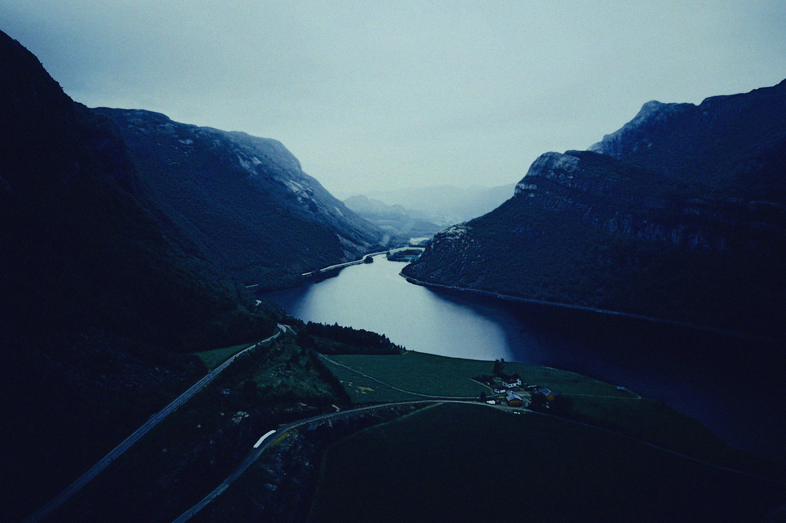 Aerial view of a fjord with mountains on either side and a small road winding through green fields at the bottom.