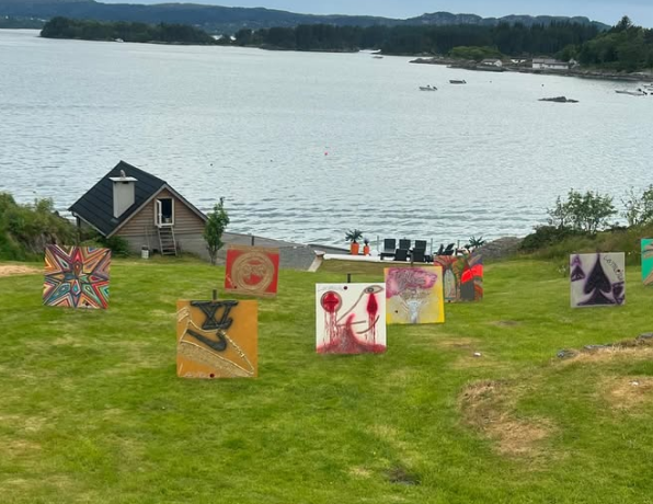 Paintings displayed on the grass near a lake with a small wooden shed and outdoor seating area.