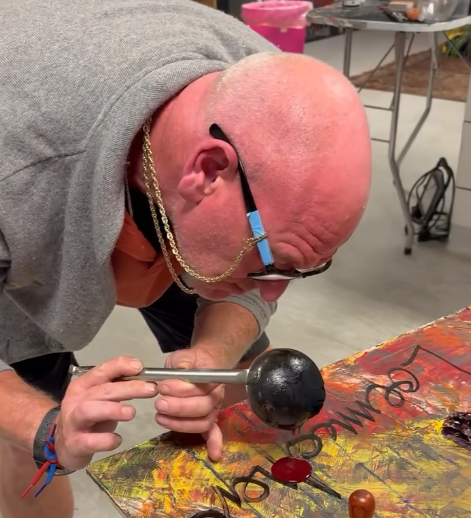 A bald man with glasses and a chain necklace is holding a black object with a metal rod, bending over a table covered with colorful paint or artwork, in what appears to be an art studio or workshop.