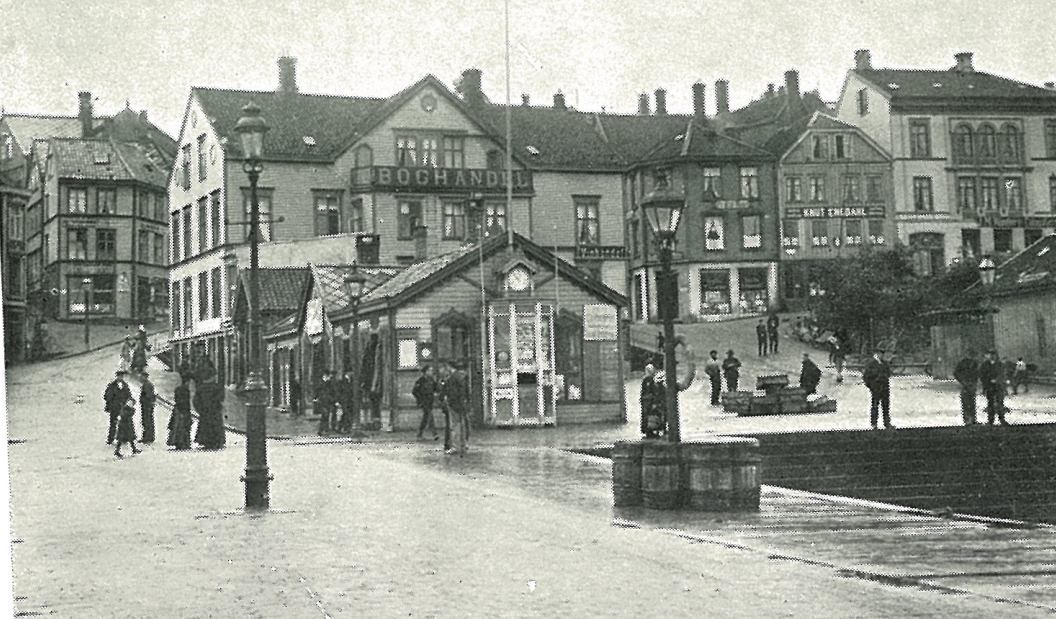 An old black and white photo of a seaside town with buildings, street lamps, and people walking around, mainly in front of a small shop named 'BOGHANDL'.