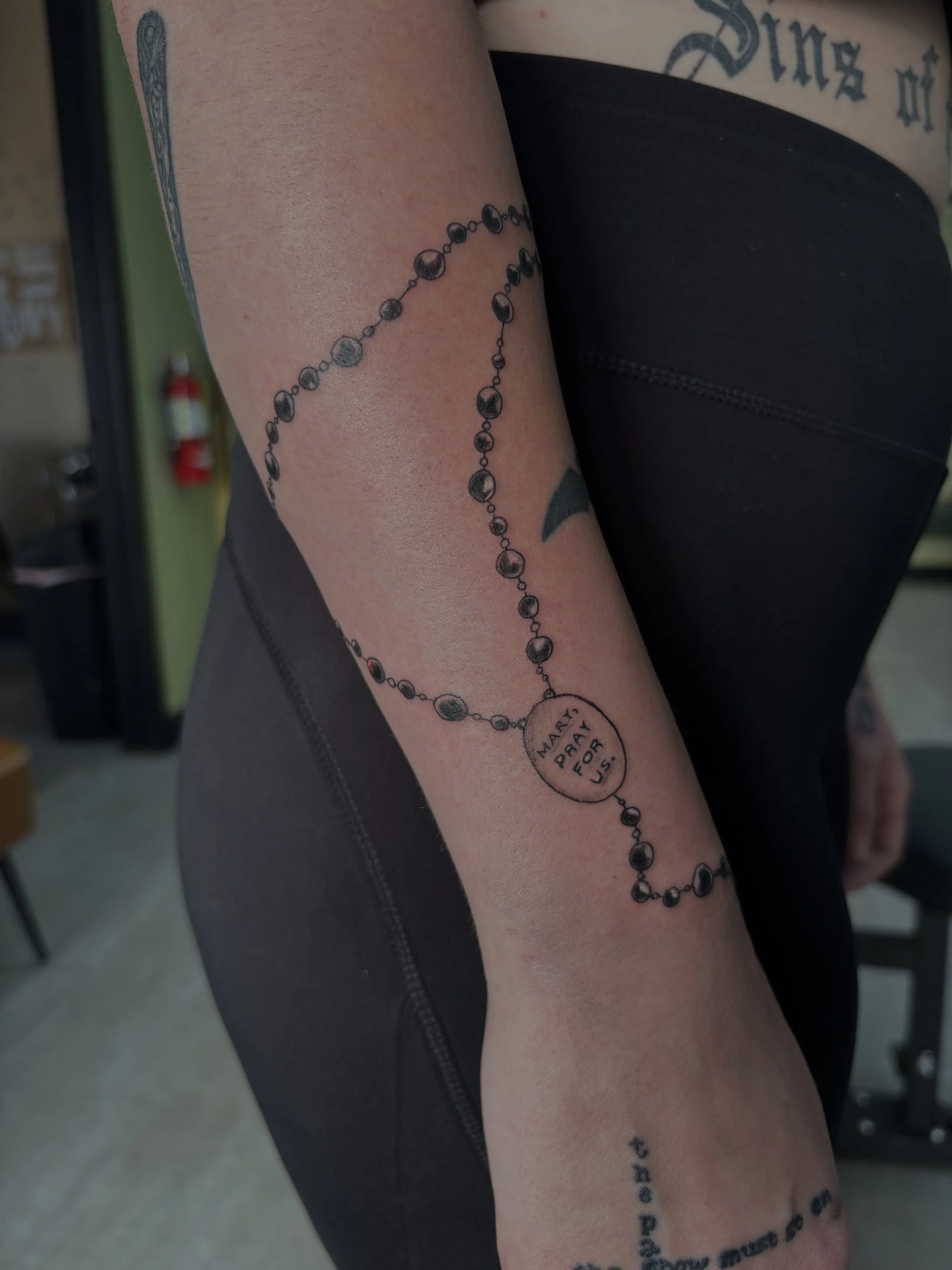 Tattoo of a rosary with a circular medallion reading 'MARY PRAY FOR U.S.' on a person's forearm.