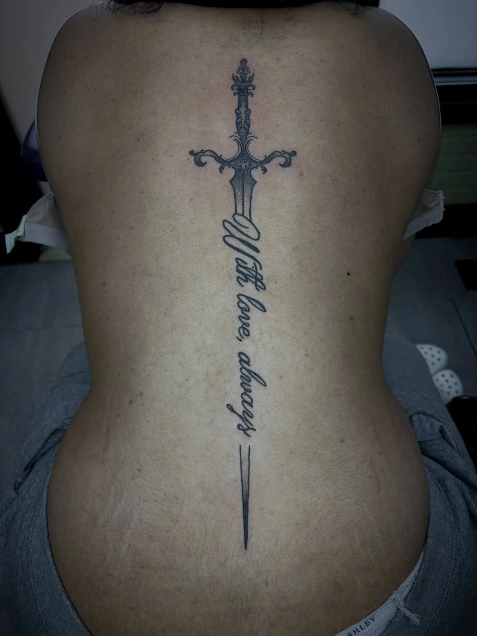A tattoo of a sword with a detailed hilt and a long blade, running vertically down a person's back. The sword's blade intersects with a cursive script that reads "You love always."