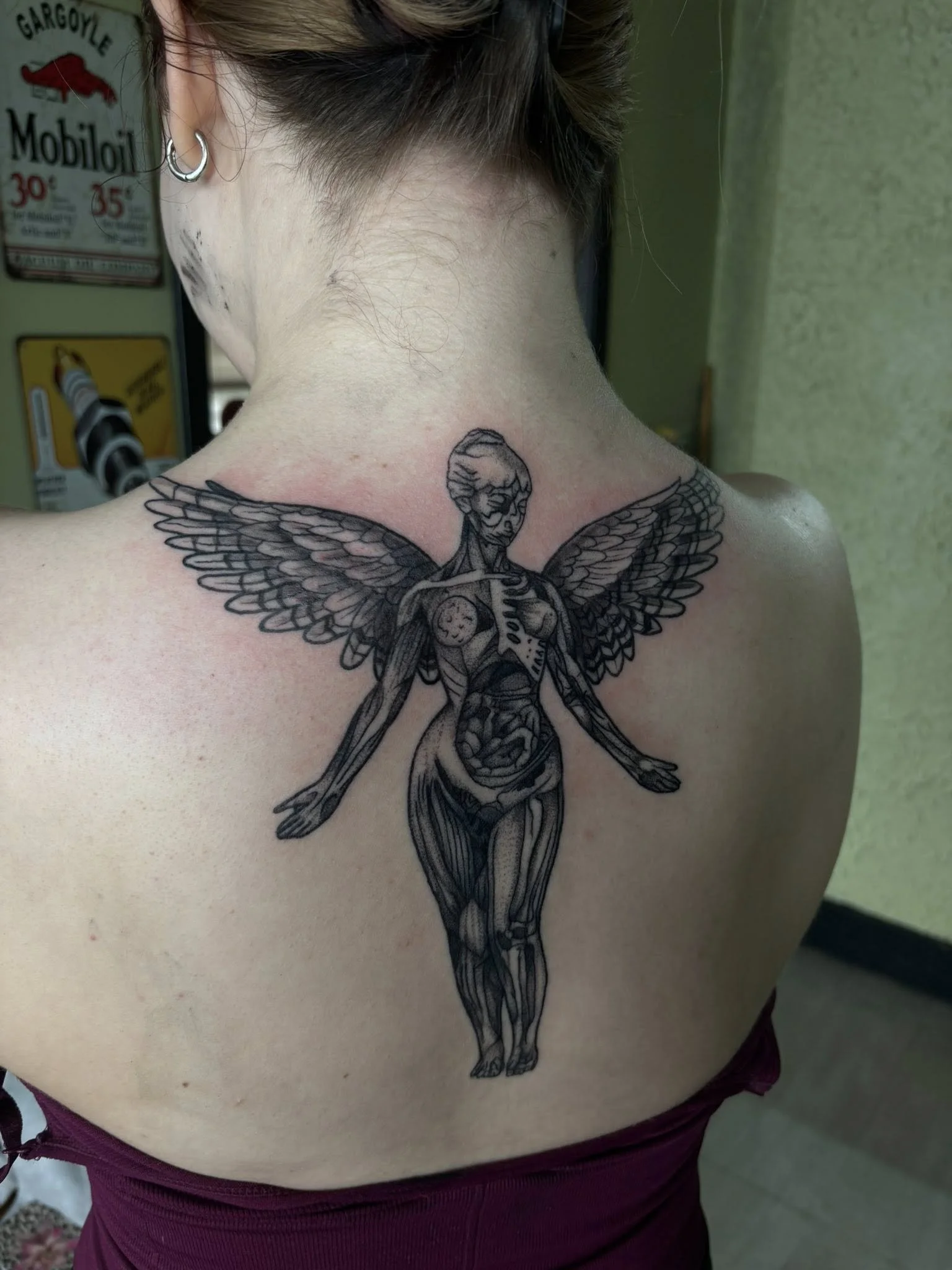 A large tattoo on a person's back featuring a humanoid figure with outstretched wings, detailed muscles, and internal organs visible, rendered in black ink with fine line work.