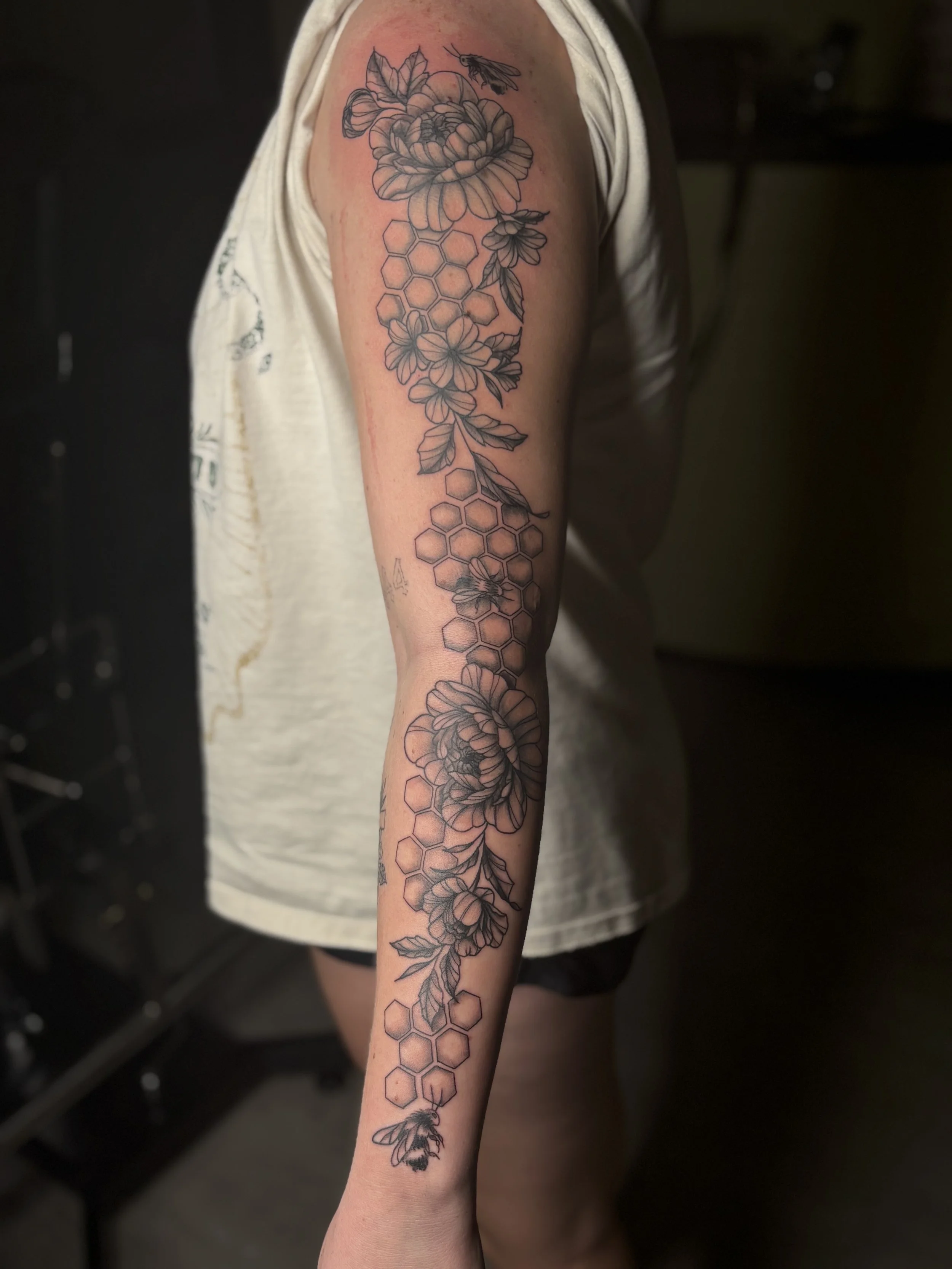 Line art tattoo on person's upper arm featuring flowers, honeycomb patterns, and bees.