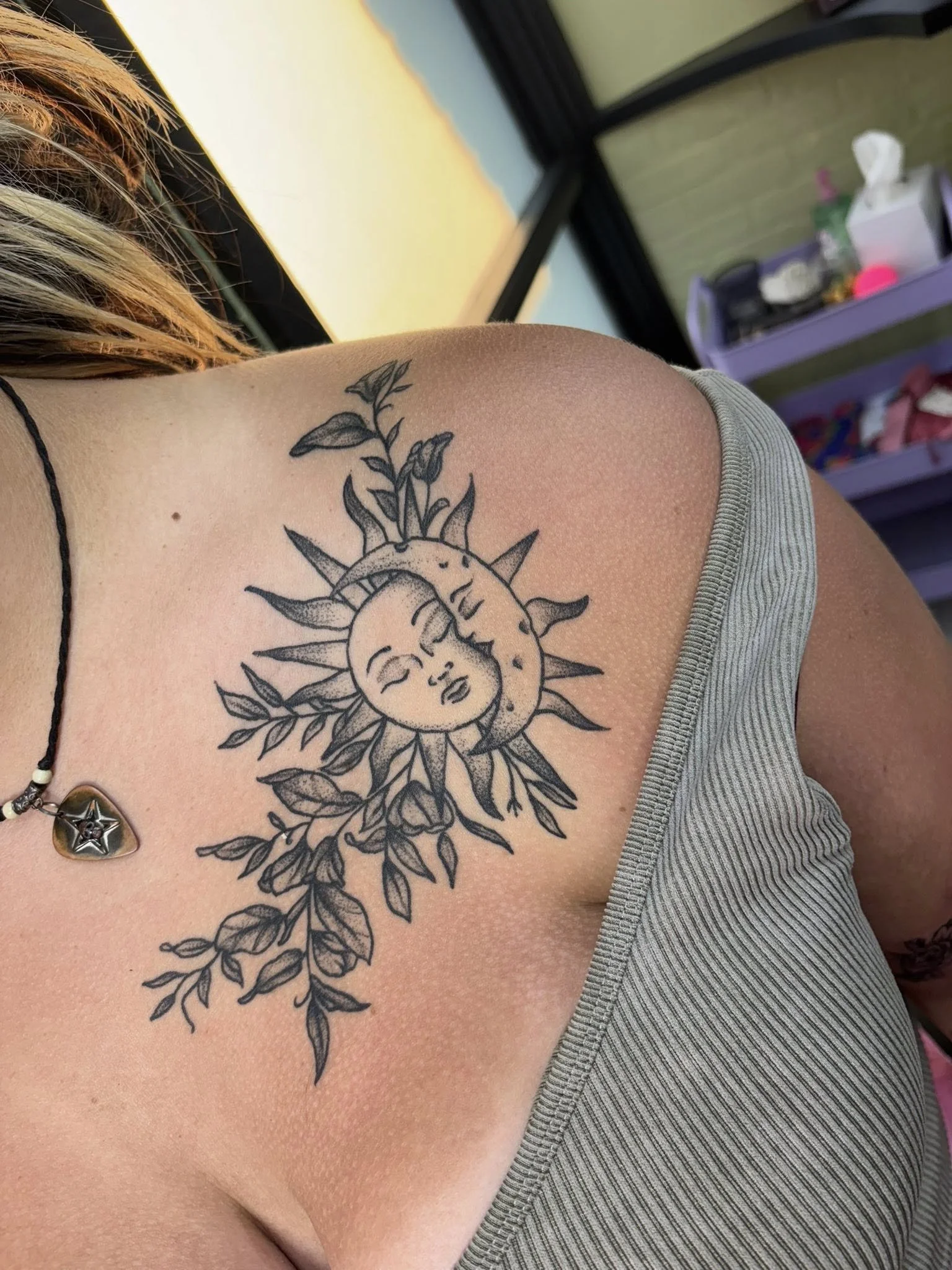 Tattoo of a sun and moon with a face, surrounded by leaves and branches, on a person's shoulder.