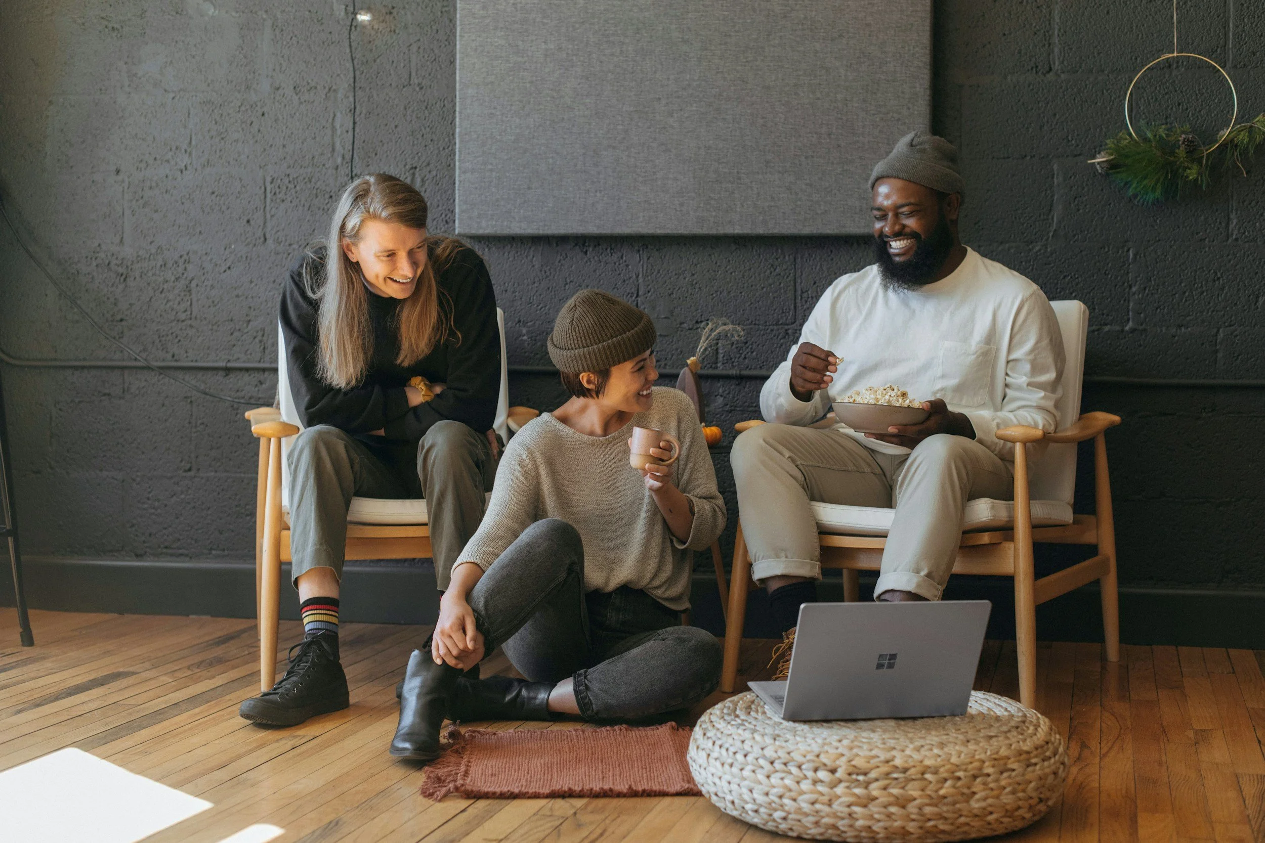 Four friends in a cozy living room, sitting and chatting while having snacks and drinks, with a laptop on a woven pouf in front of them.