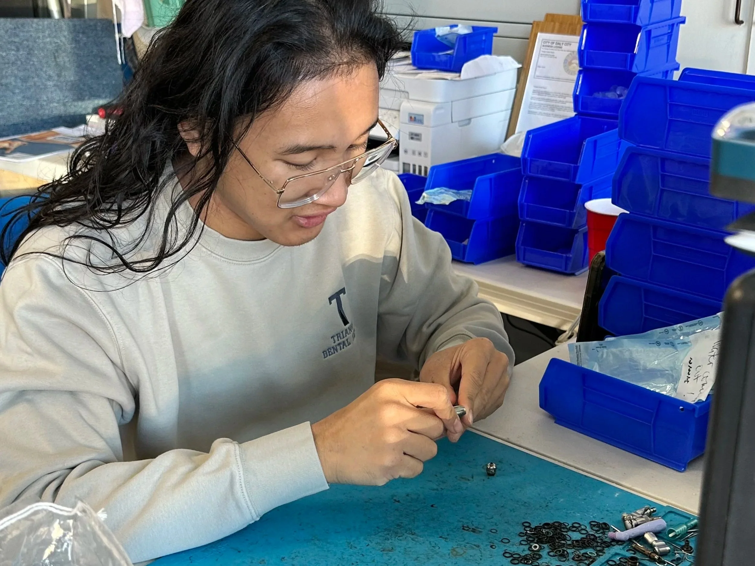 A woman with long dark hair, glasses, and a beige sweatshirt is sitting at a table, working on small mechanical components with small tools. There are blue storage bins, papers, and equipment in the background.