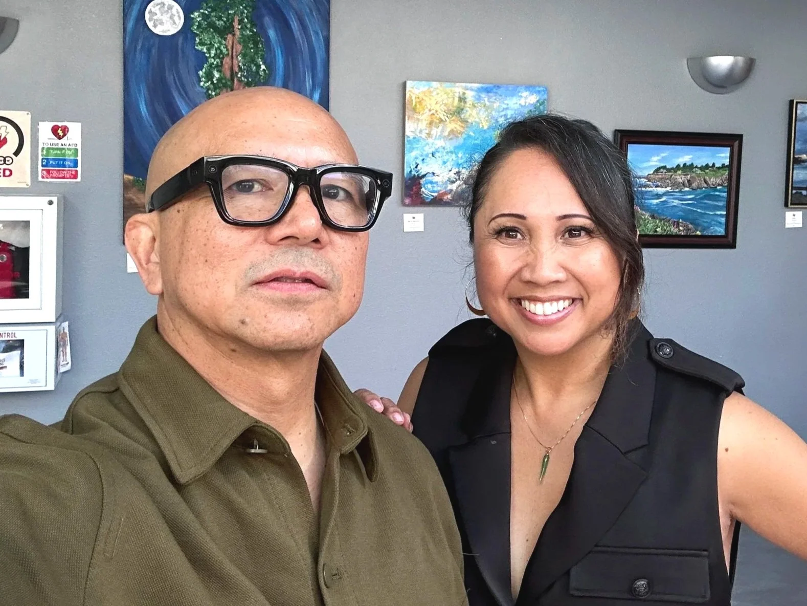 A man and woman taking a selfie inside an art gallery. The man is bald, wearing glasses and a brown shirt, while the woman has dark hair, is smiling, and is dressed in a black sleeveless top. The background includes colorful landscape paintings and artwork on the wall.