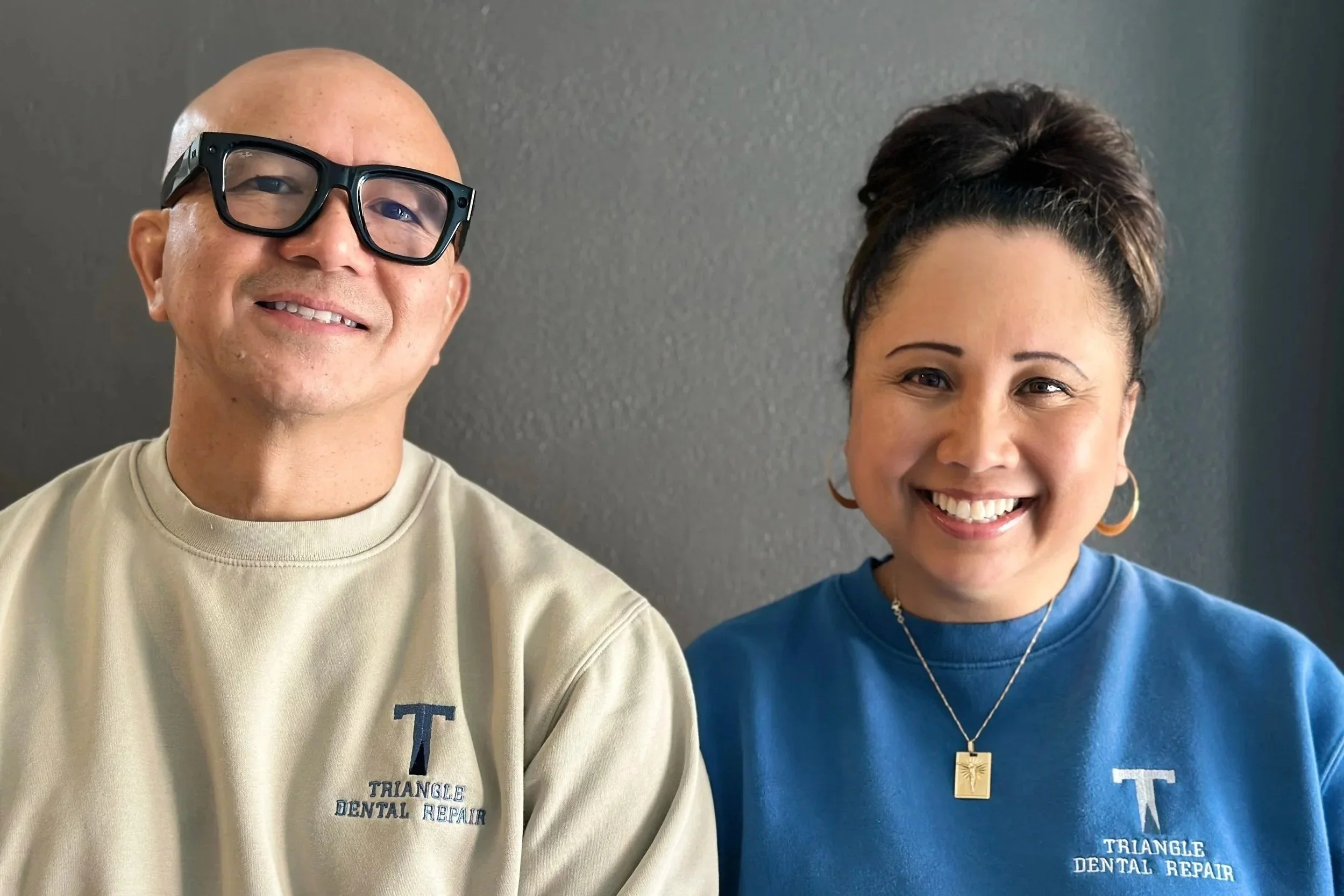 A man and woman smiling for a selfie against a gray background, both wearing Triangle Dental Repair shirts.