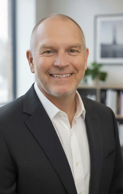 A smiling man dressed in a black suit and white shirt standing in an office, with a bookshelf and a potted plant in the background.