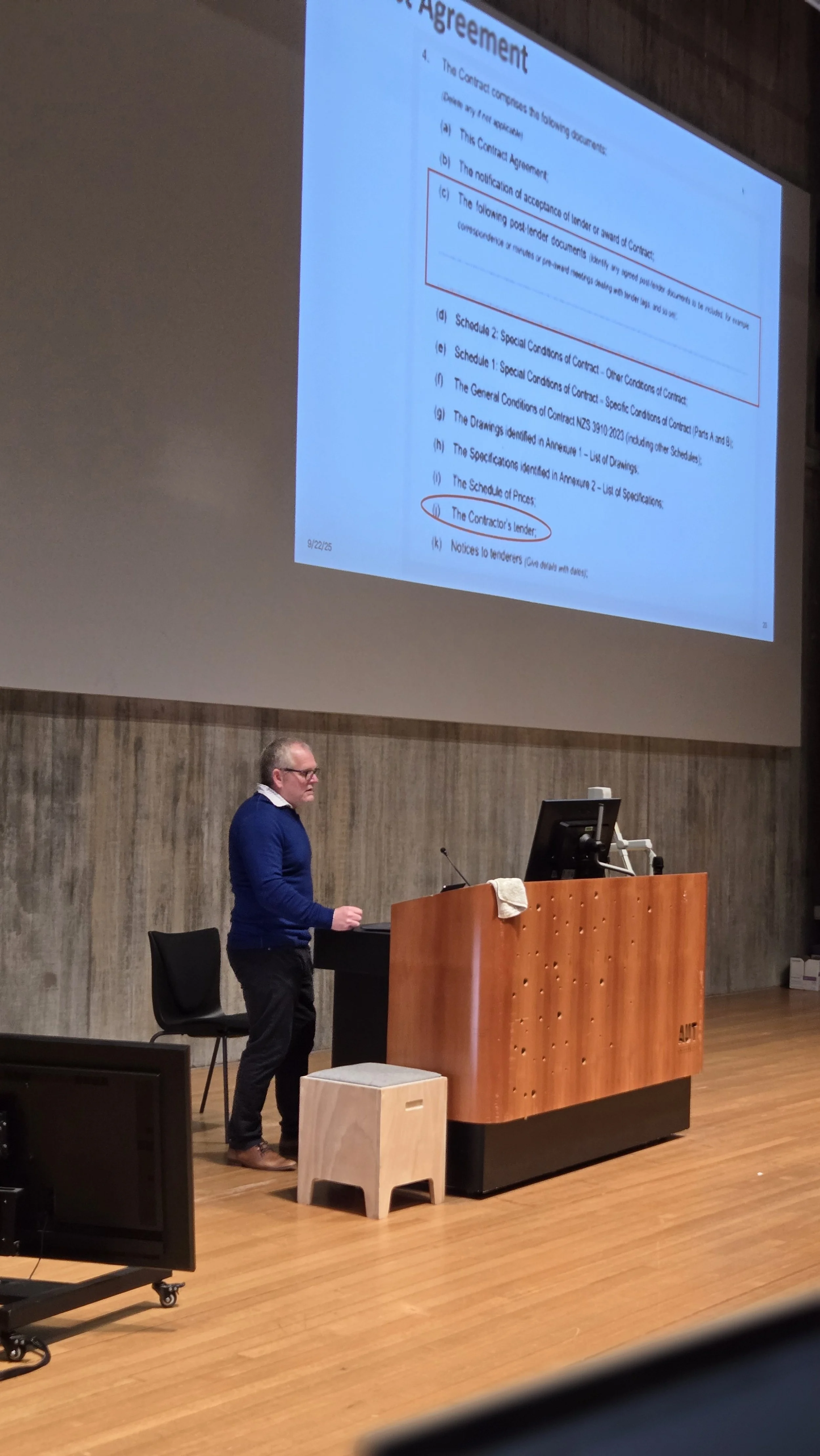 A man in glasses and a blue sweater standing at a wooden podium in a lecture hall, giving a presentation displayed on a large screen behind him.