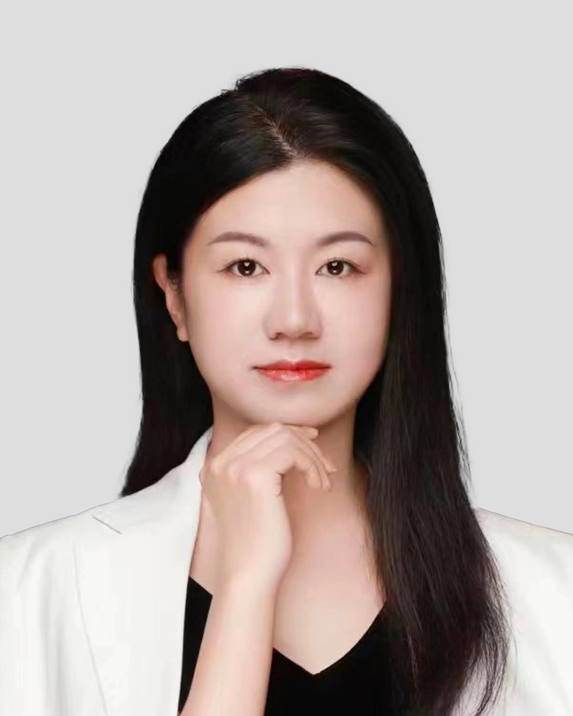 A woman with long black hair, wearing a white blazer and black top, posing with her chin resting on her hand against a gray background.