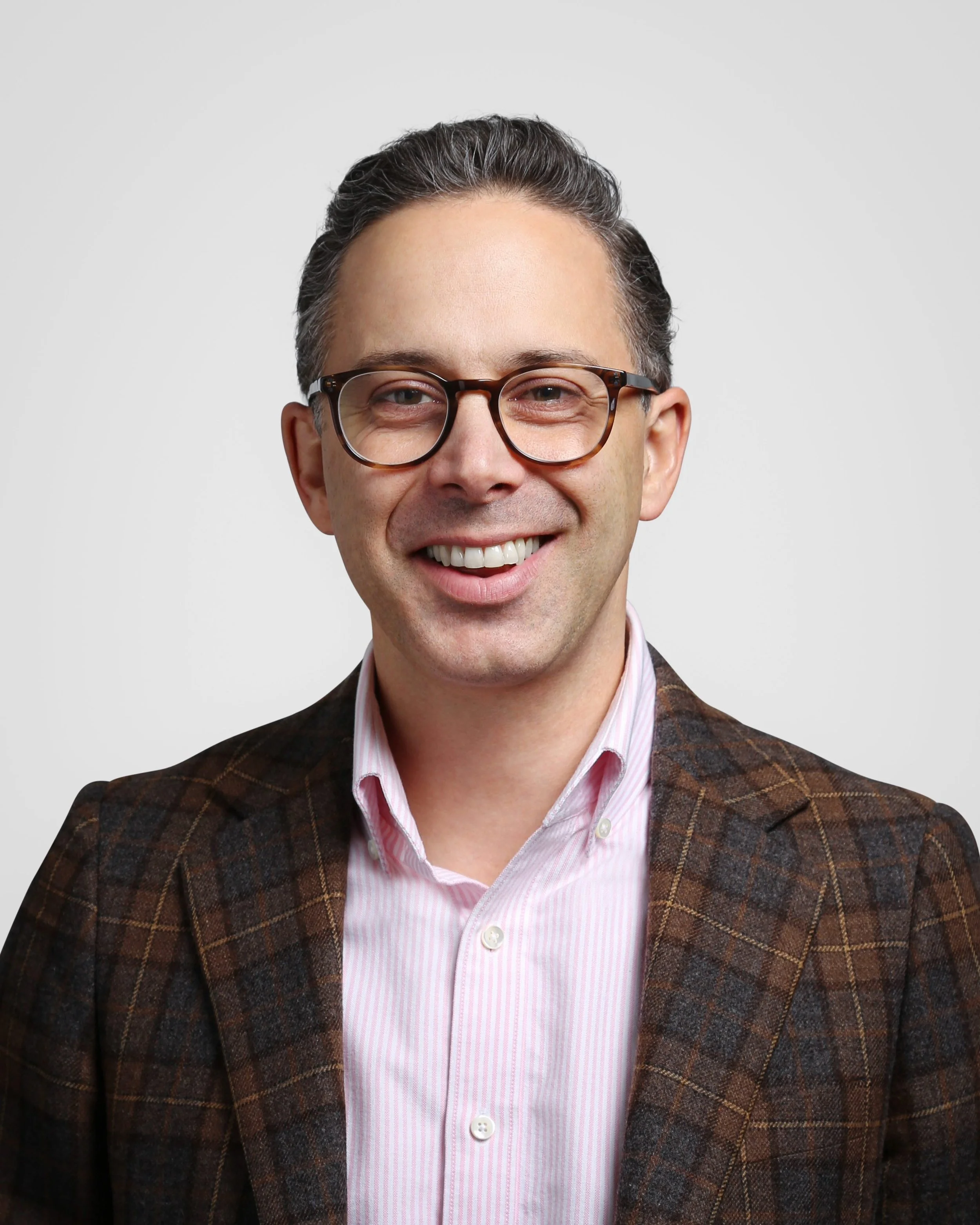 A man with dark hair, glasses, and a light pink collared shirt smiling, wearing a brown plaid jacket.