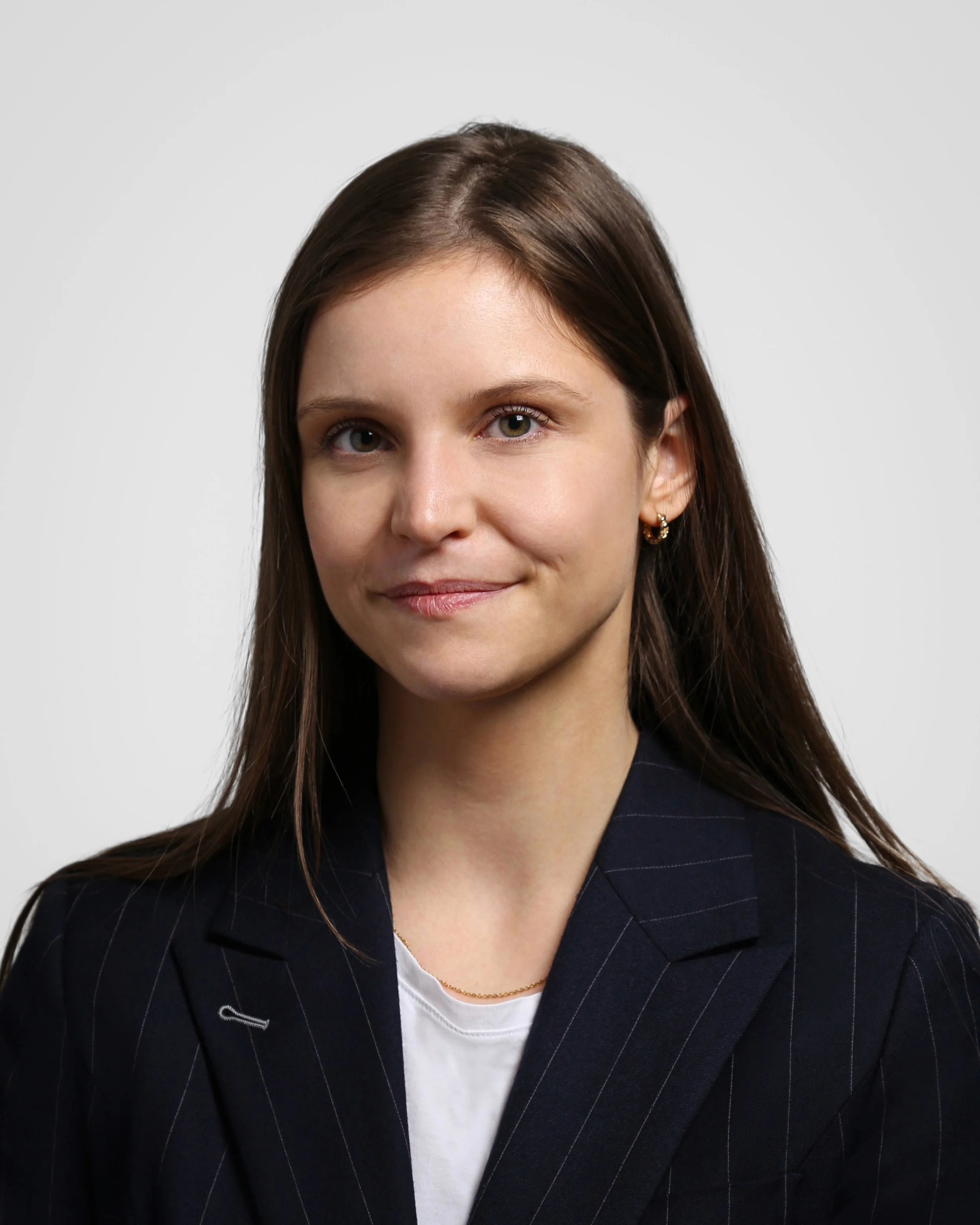 A woman with straight brown hair wearing a dark pinstripe blazer and a white shirt, smiling softly against a plain background.