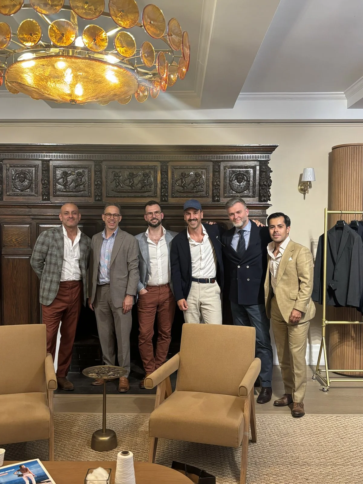 A group of seven men in formal and semi-formal attire standing together in a well-decorated room, smiling at the camera. There are armchairs, a small table, and clothing rack in the background.