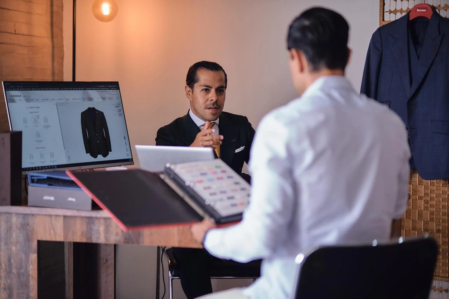 Two men in a clothing store discussing a suit, one sitting behind a counter with a computer screen showing the suit, and the other facing him holding a color palette, with jackets hanging on the wall behind.