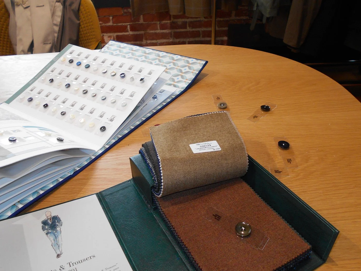 A wooden table displaying fabric swatches, buttons, and catalogs for suit and trousers selection in a store.