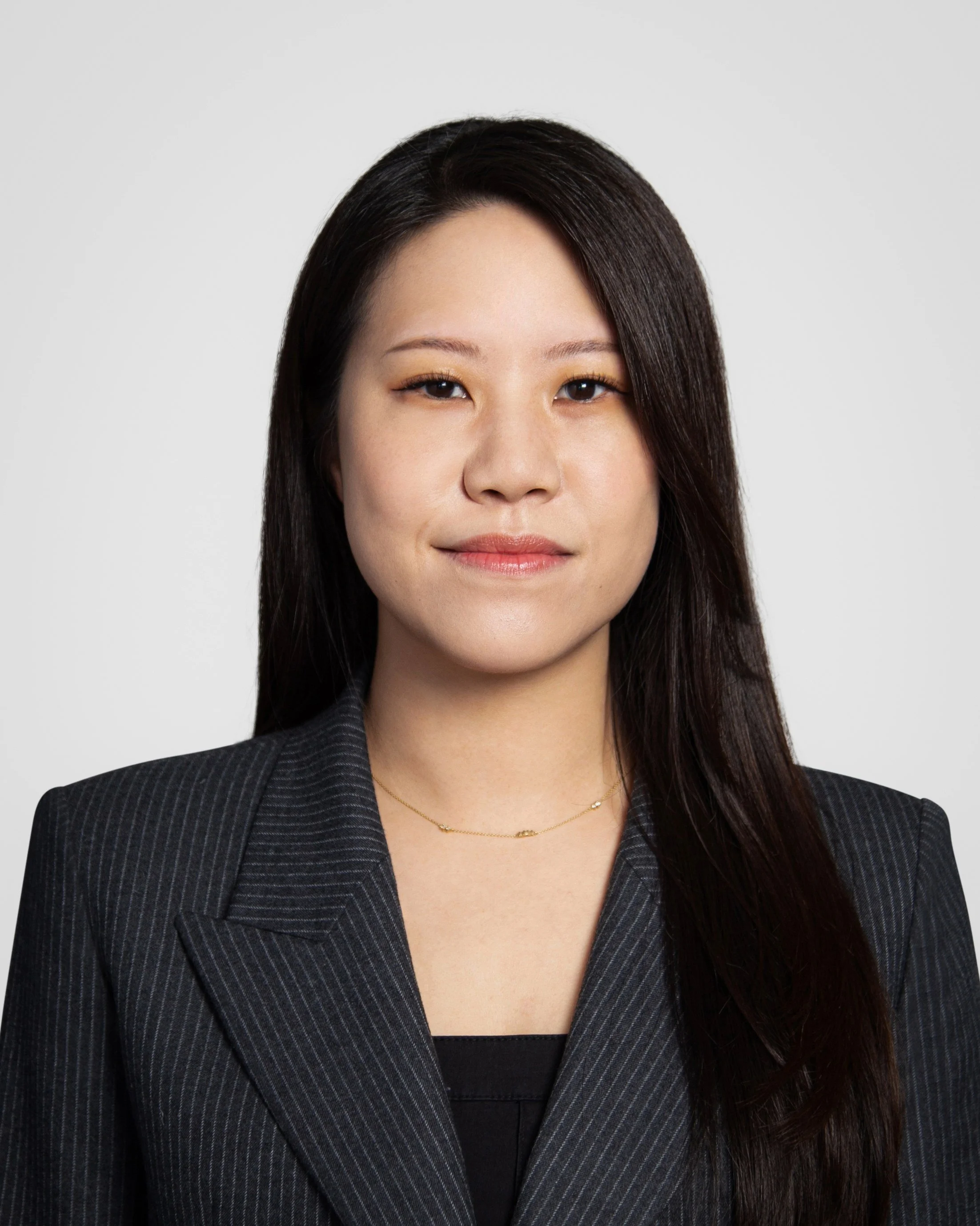Professional portrait of a young Asian woman with long dark hair, wearing a black pinstripe blazer and a delicate gold necklace, against a plain light gray background.