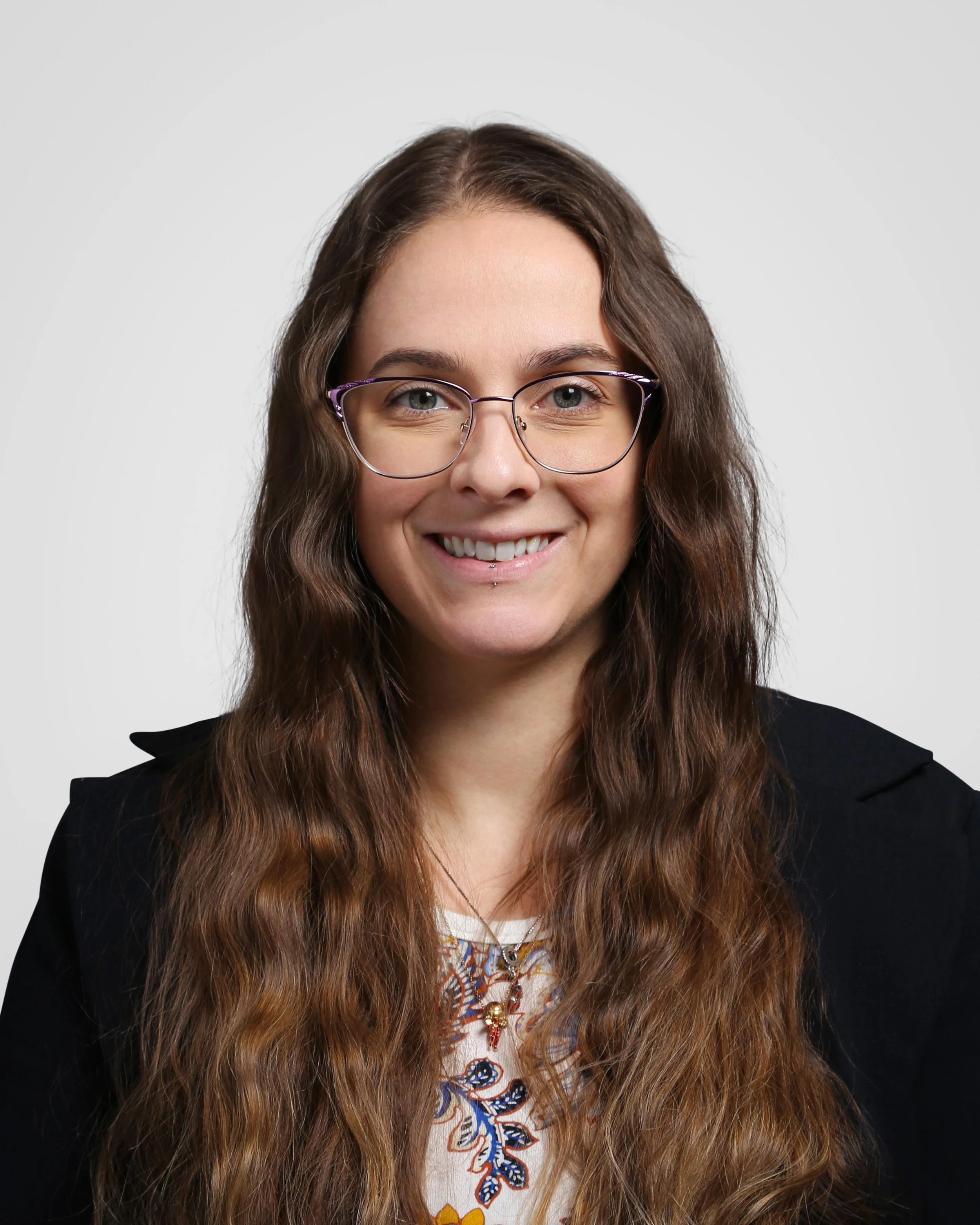 Headshot of a woman with long wavy brown hair, wearing glasses, a black blazer, and a patterned blouse, smiling at the camera against a plain light gray background.