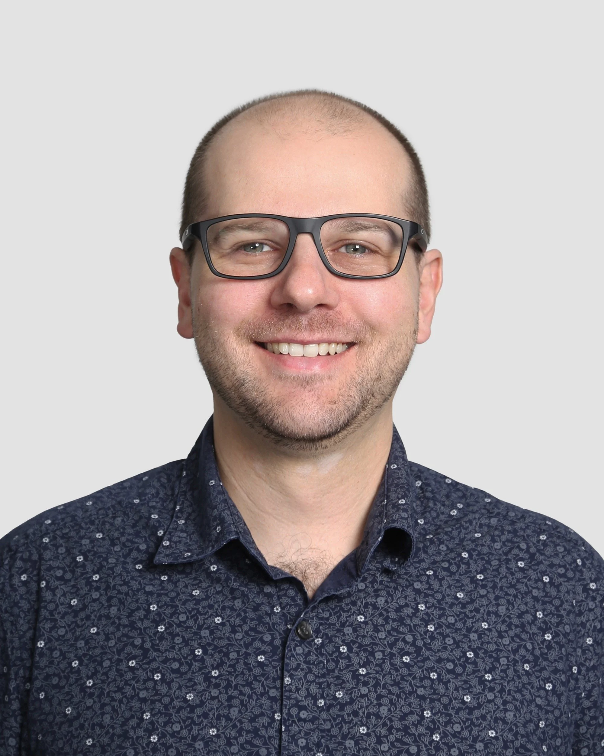 A smiling man wearing black-rimmed glasses and a navy blue button-up shirt with floral patterns against a light gray background.