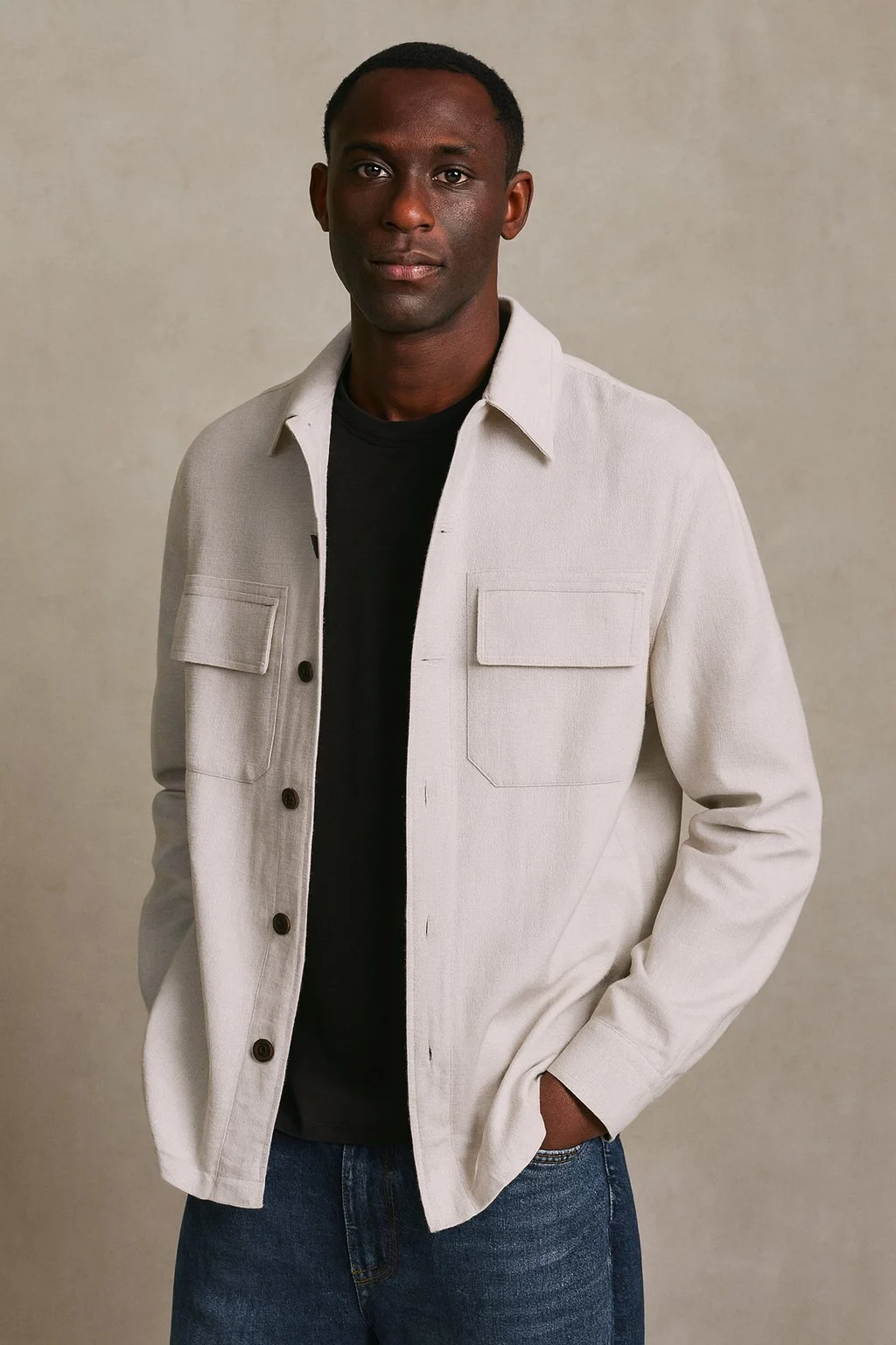 A man standing against a plain background, wearing a cream-colored jacket over a black shirt and blue jeans.