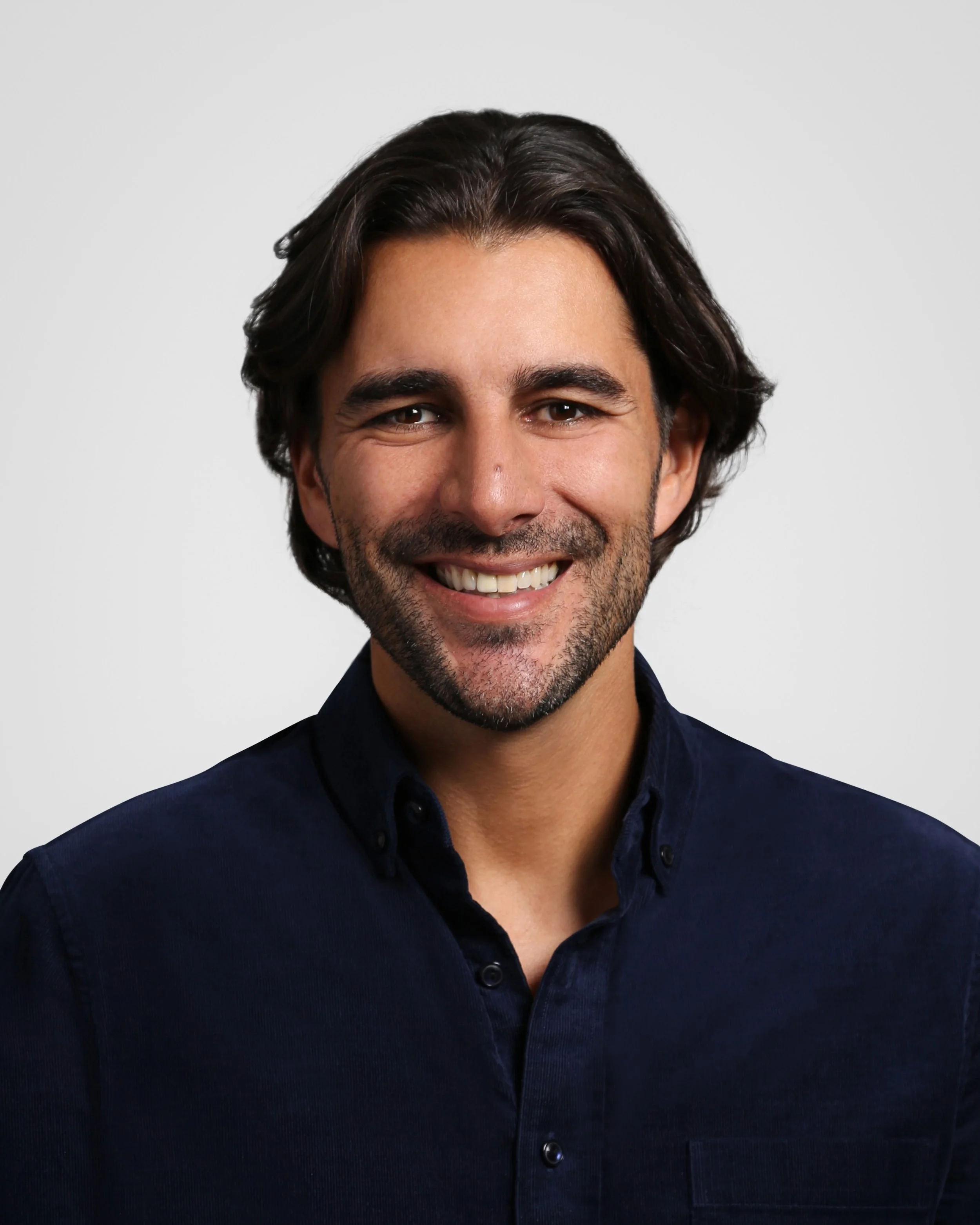 Smiling man with dark hair, beard, wearing a dark button-down shirt, against a plain light gray background.