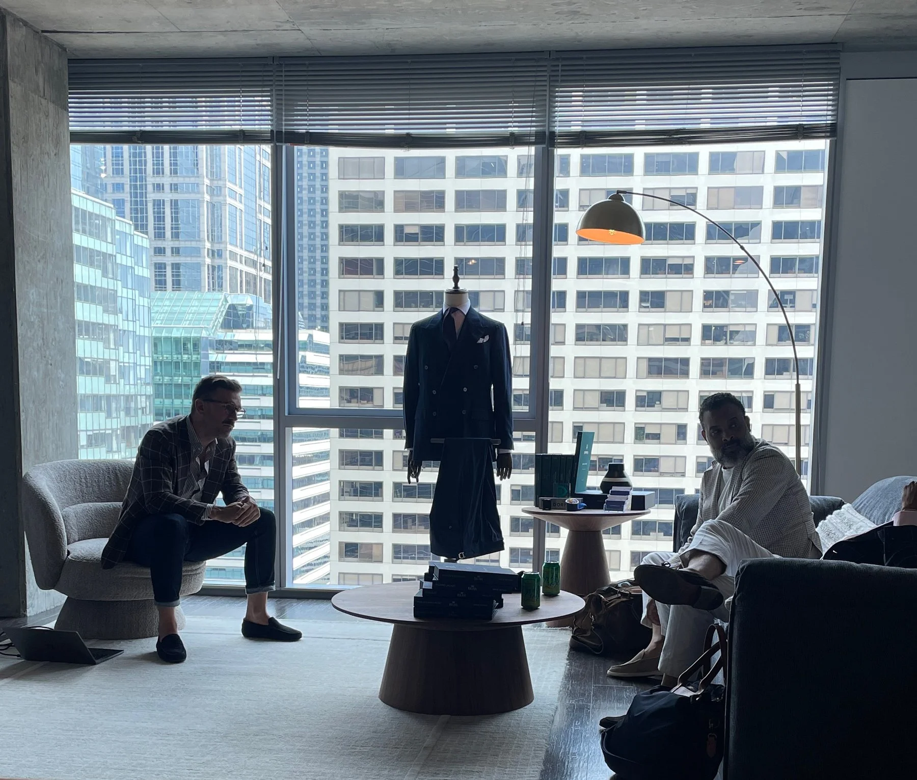 Two men sitting in a modern office with large windows, one wearing casual attire and the other in a light-colored outfit, with a mannequin dressed in a suit and tie in the center of the room.