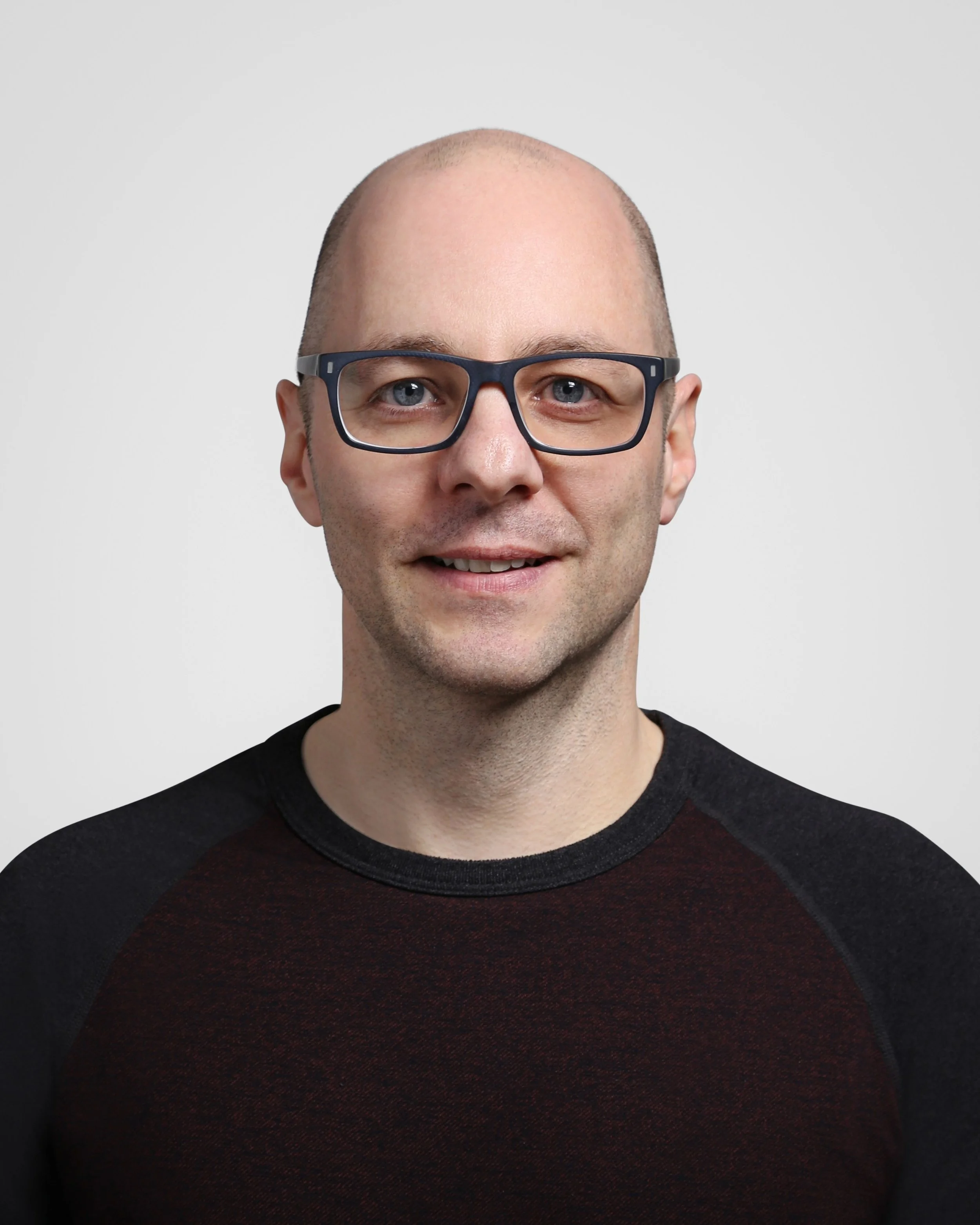 Portrait of a bald man with glasses, smiling, wearing a black and dark red shirt against a plain light gray background.