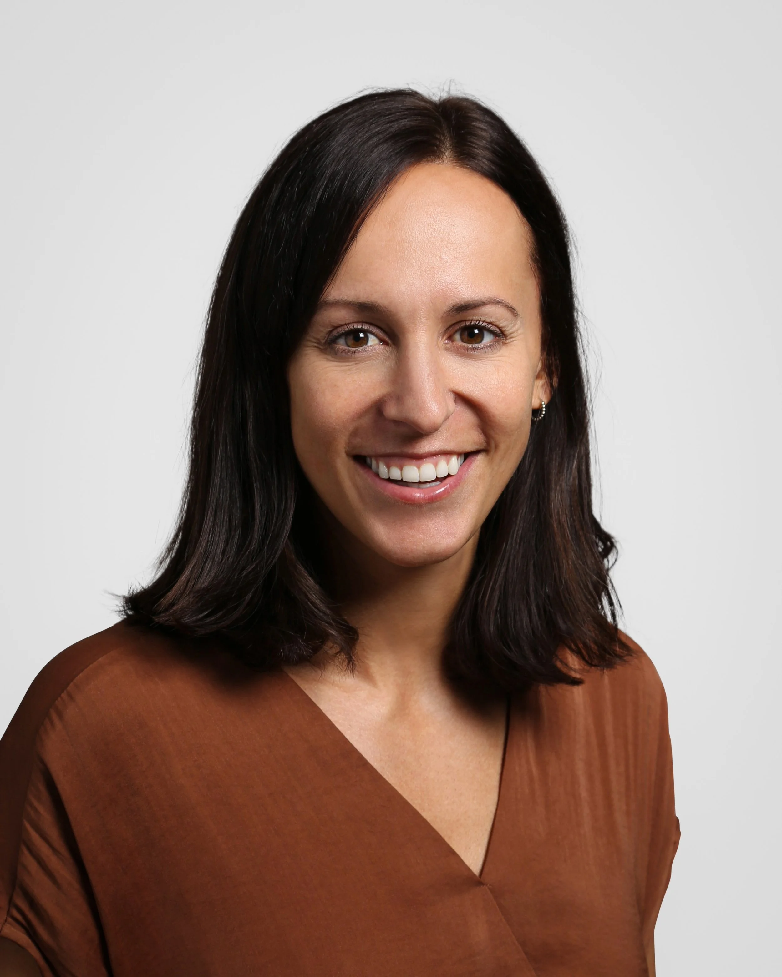 Headshot of a smiling woman with shoulder-length dark hair, wearing a brown top, against a plain white background.