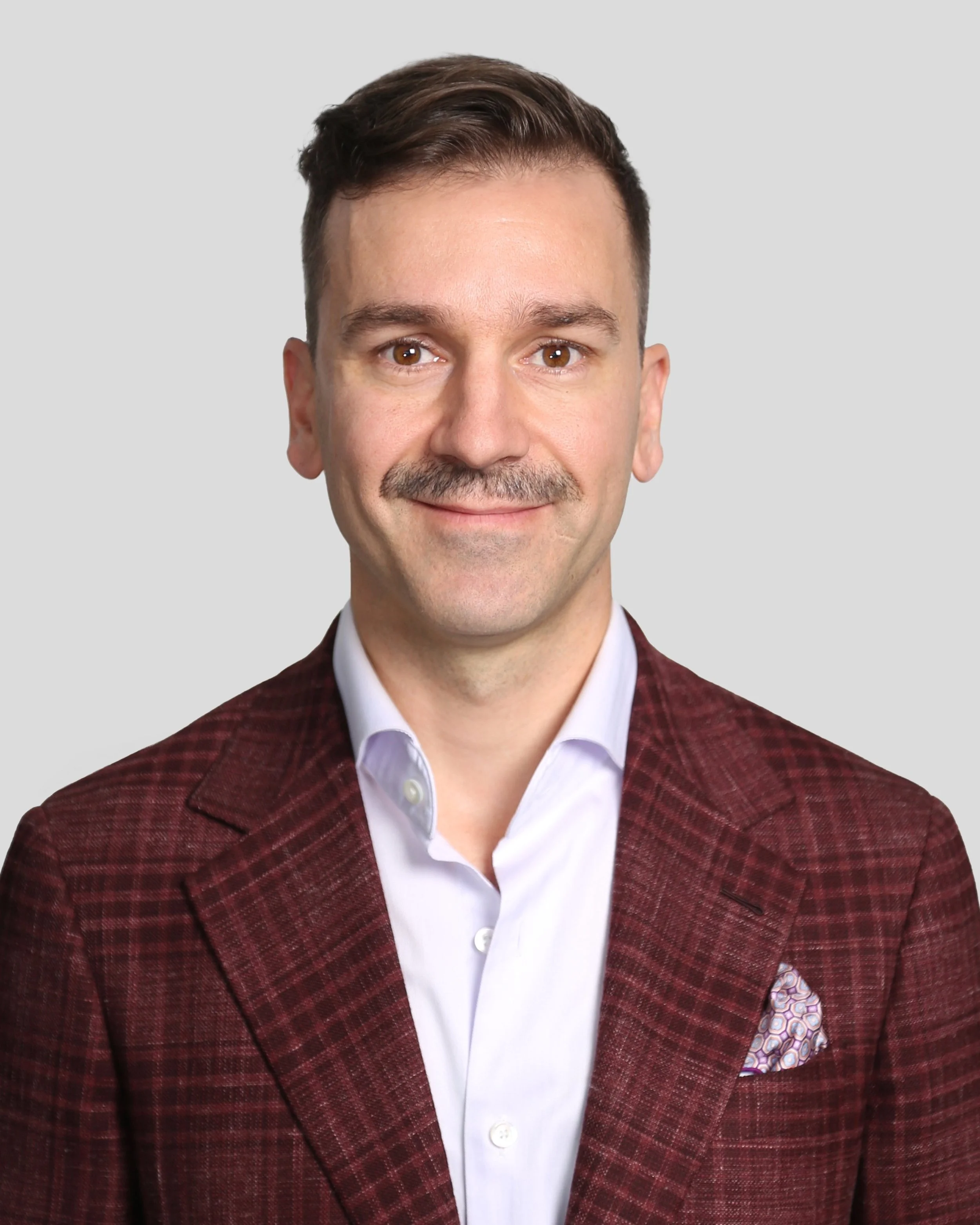A man with short brown hair, a mustache, wearing a white collared shirt and a maroon checkered blazer, smiling against a plain light gray background.