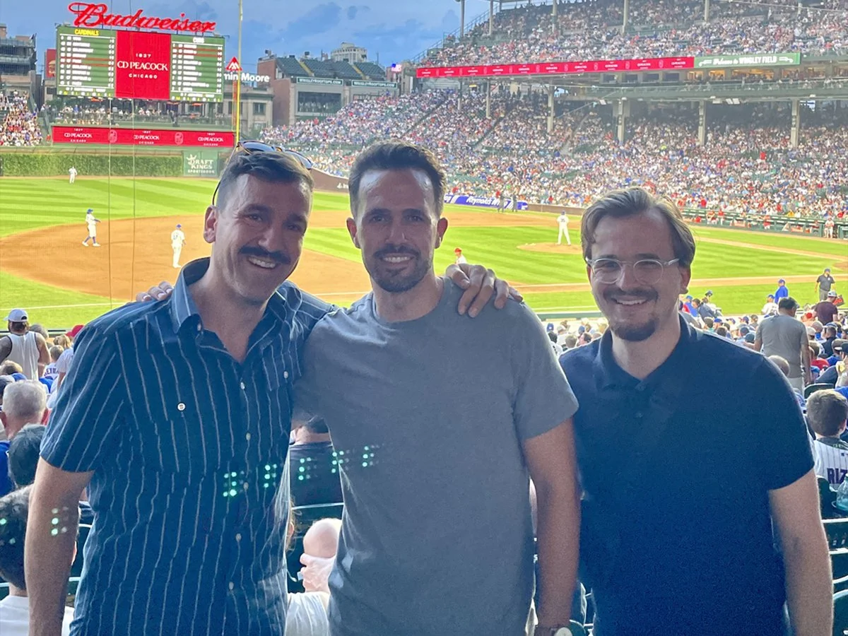 Three men smiling at a baseball game in a stadium, with the field and crowd visible in the background.