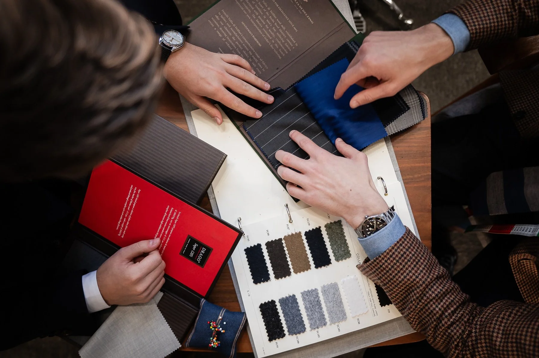 Two people examining fabric swatches at a table with a sample booklet, fabric samples, and color samples for upholstery or clothing design.