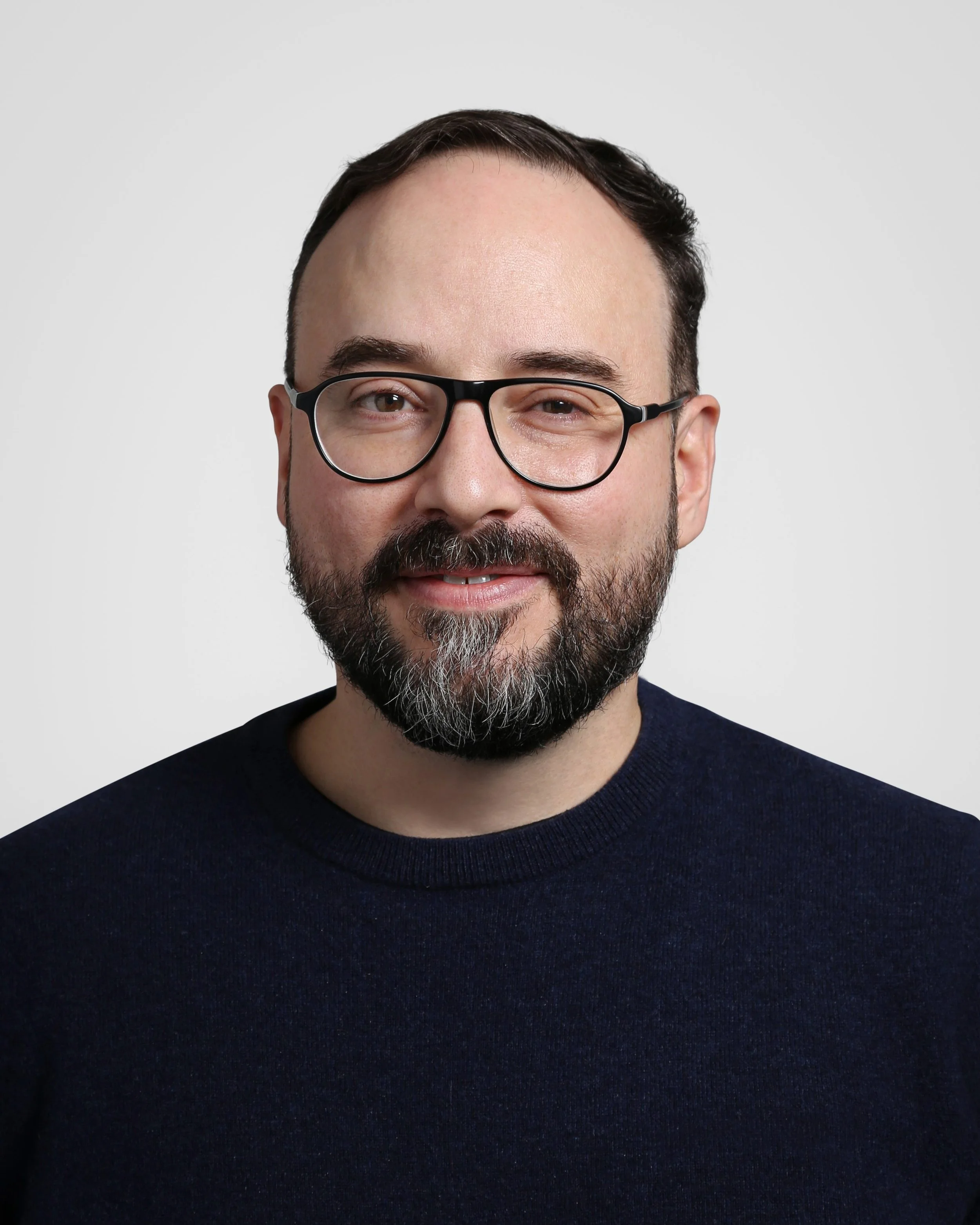 A man with glasses, a beard, and dark hair smiling against a plain light background.
