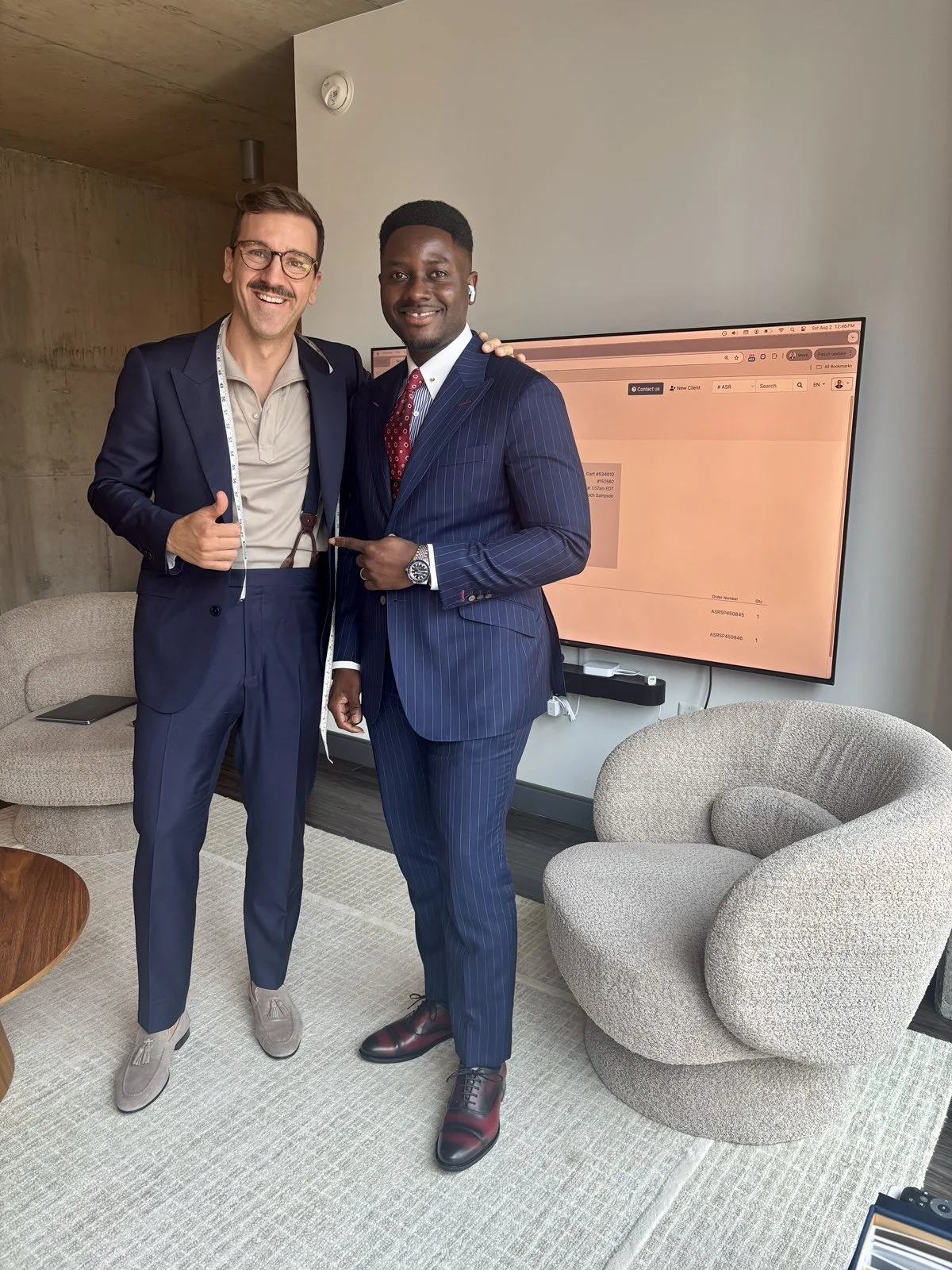 Two men in suits standing in a modern office, smiling and posing for the photo. One man is wearing glasses and a navy suit, the other is wearing a pinstripe suit and pointing at the first man. They are in front of a large TV screen and modern chairs.