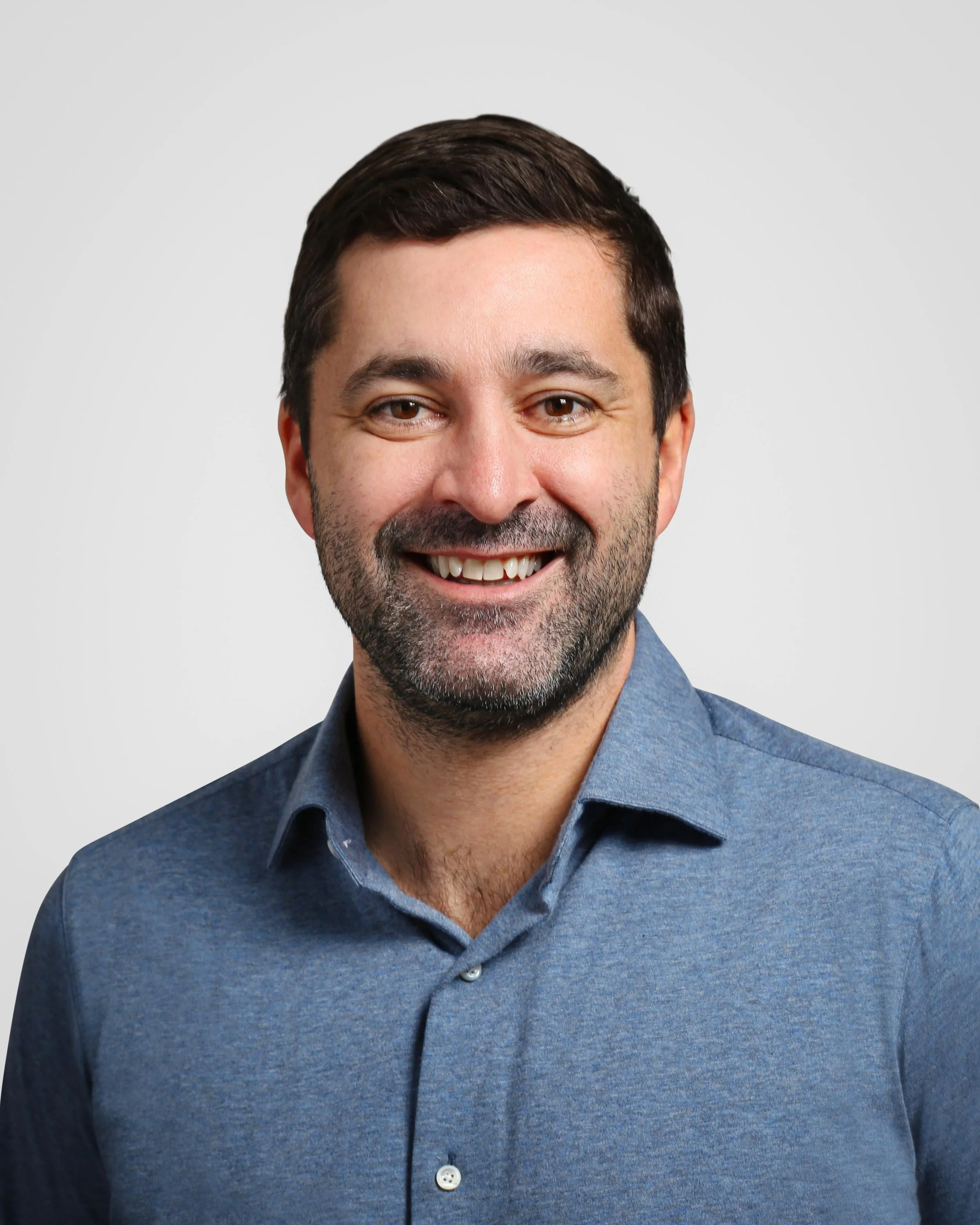 A smiling man with dark hair and a beard, wearing a blue collared shirt, against a light grey background.