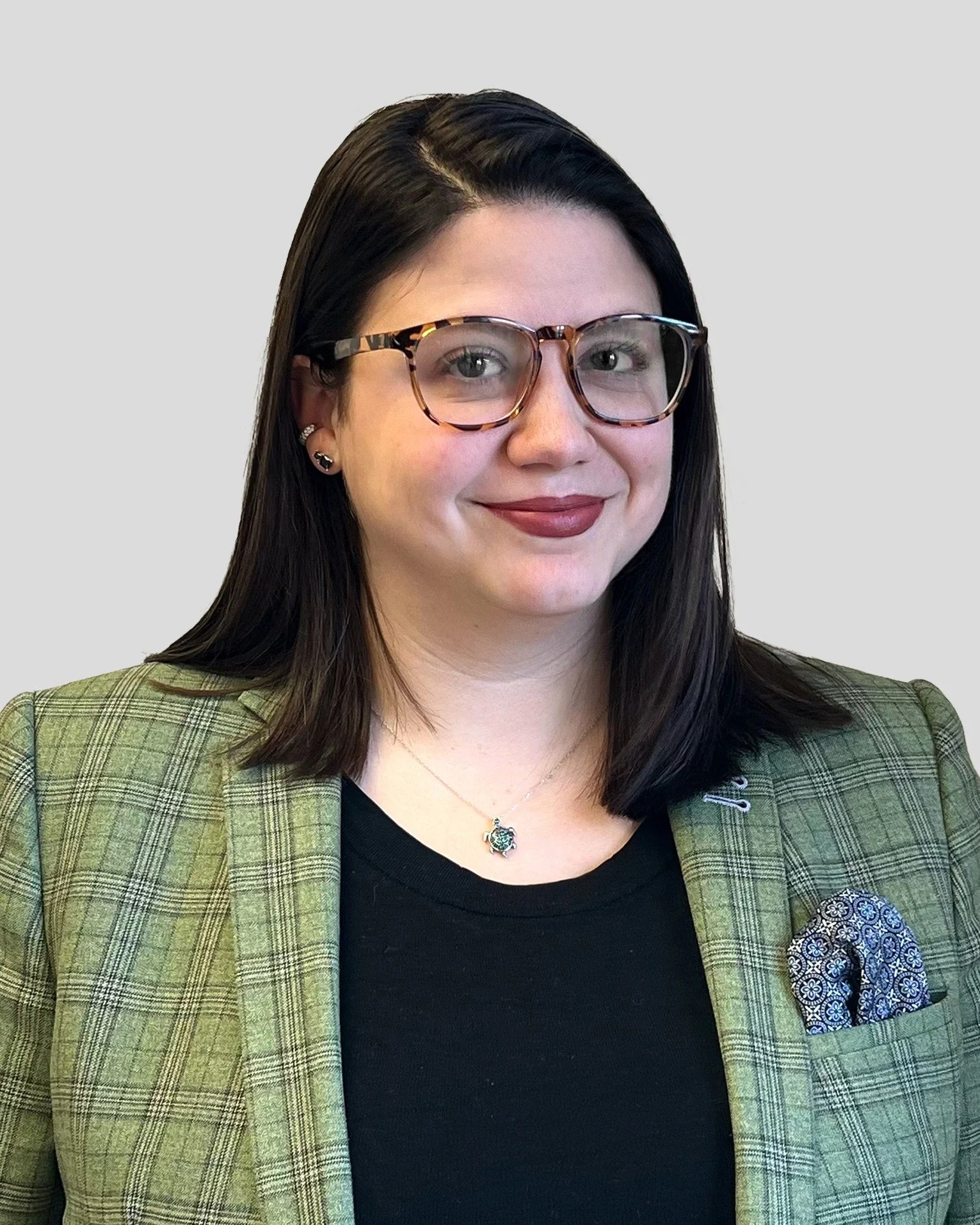A woman with dark hair, glasses, and lipstick, wearing a green plaid blazer and a black top, smiling at the camera.