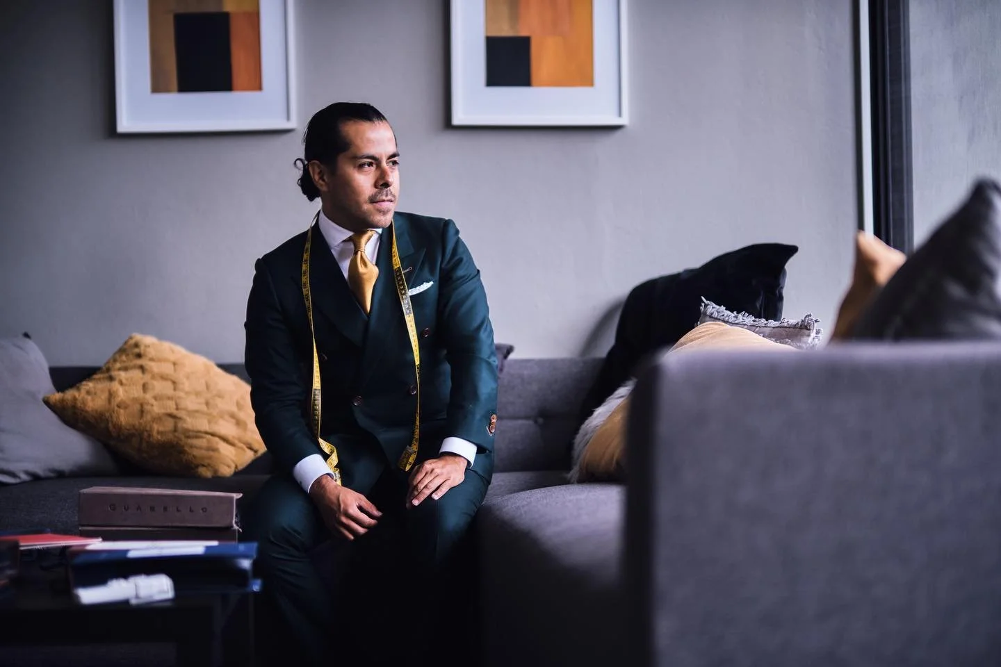 A man in a suit with a measuring tape around his neck sits on a couch, looking thoughtfully towards the right, in a modern room with framed artwork on the wall.