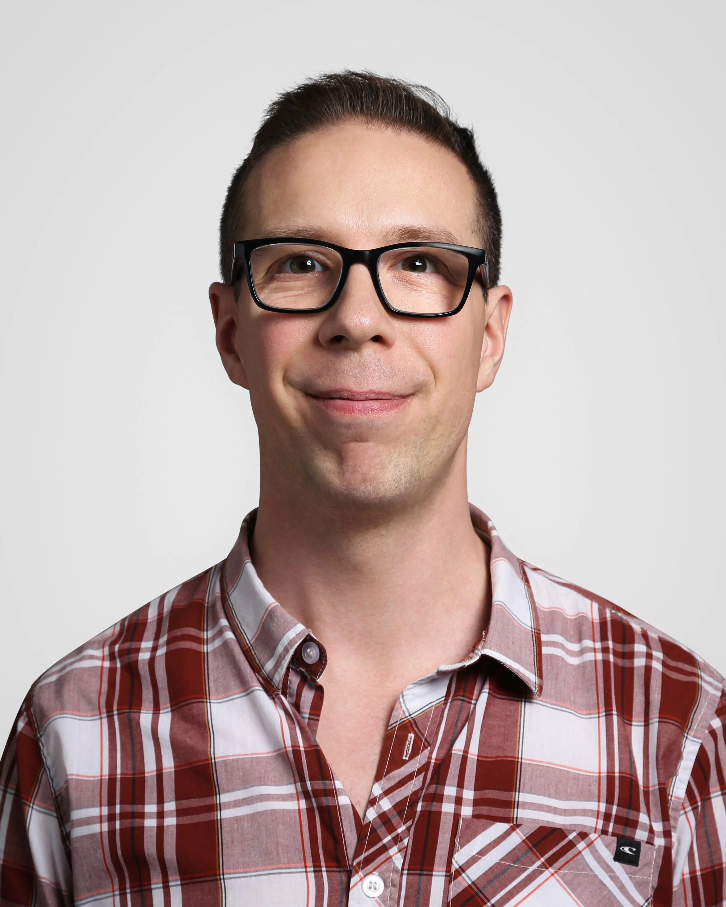 A man with short dark hair, wearing black glasses and a red plaid shirt, smiling at the camera against a plain light gray background.