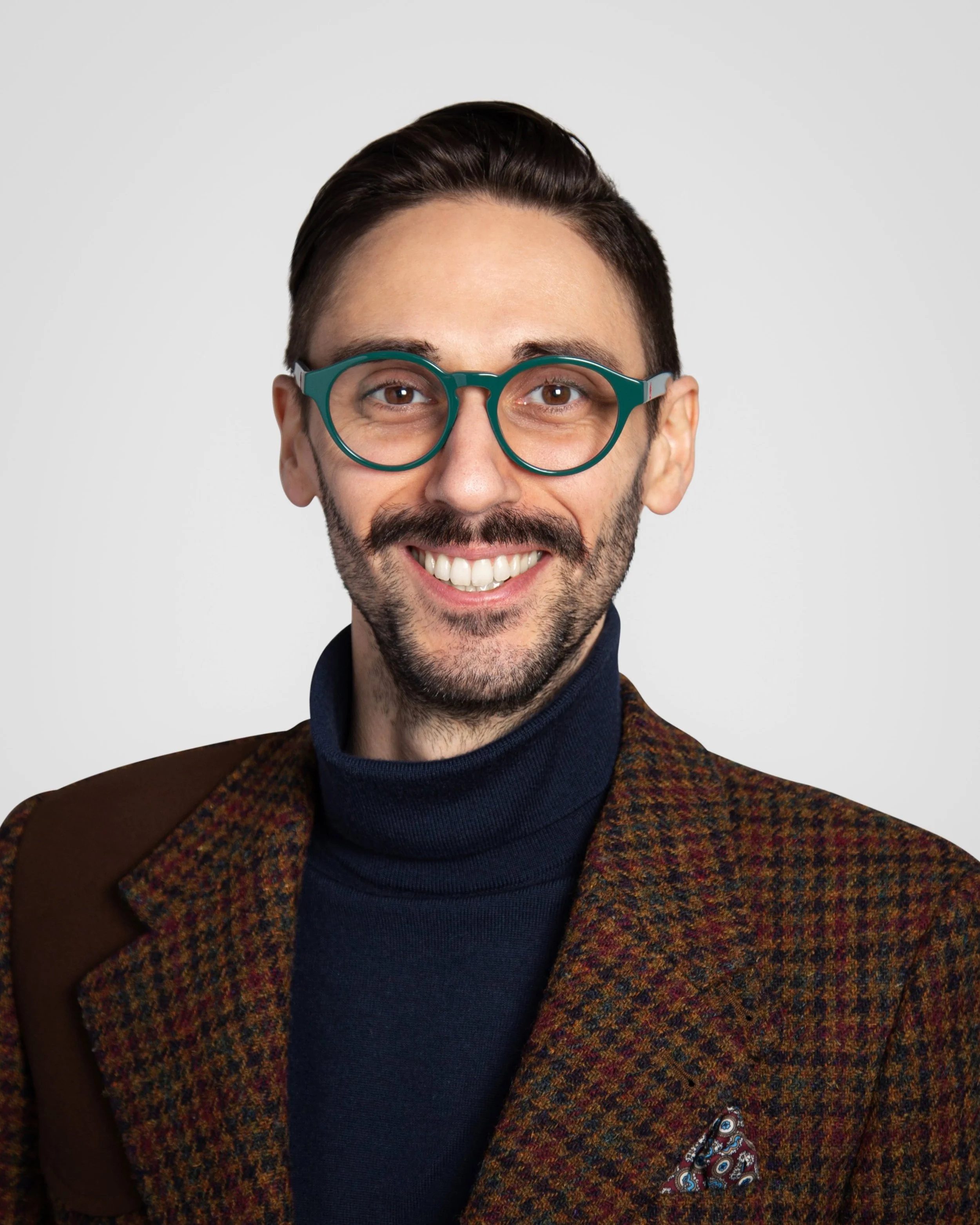 A smiling man with brown hair, wearing teal glasses, a dark turtleneck, and a plaid blazer with a pocket square.