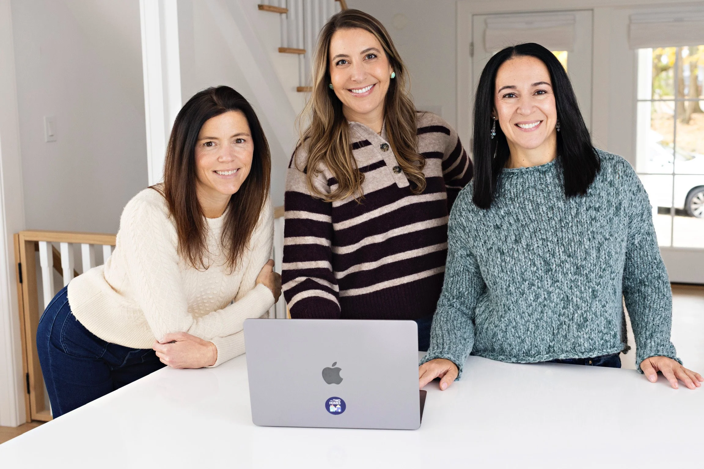 My Two Homes founders, three women, smiling and standing behind a white kitchen island with a silver MacBook laptop on it. They are indoors in a modern home, with a staircase, large windows, and an outdoor view of a car and trees in the background.
