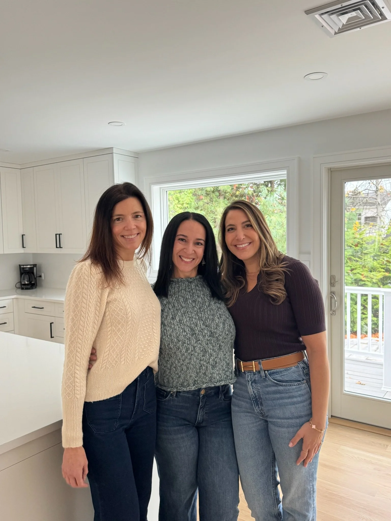Three women standing close together in a bright kitchen, smiling at the camera. There are large windows with greenery outside and a white kitchen with cabinets and a coffee maker visible in the background.