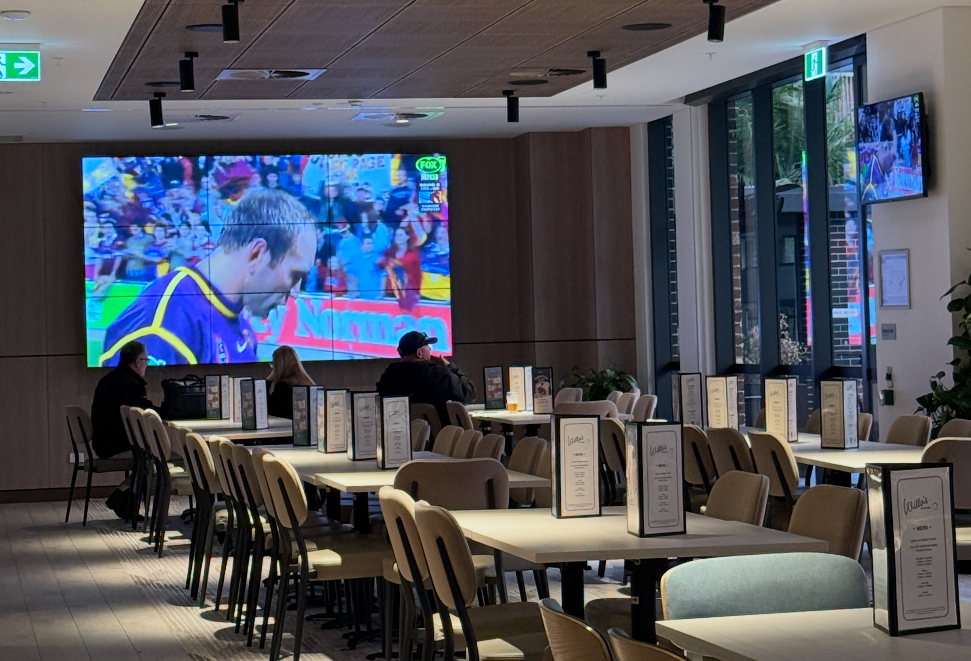 A modern indoor dining area with empty tables and chairs, a large wall-mounted TV showing a cricket match, smaller TV screens on the corners displaying the same game, and large windows letting in natural light.