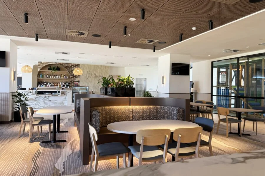 Interior of a modern restaurant or cafe with a bar area, round tables, and chairs. Decor includes plants, pendant lights, and a mix of wooden and upholstered furniture.