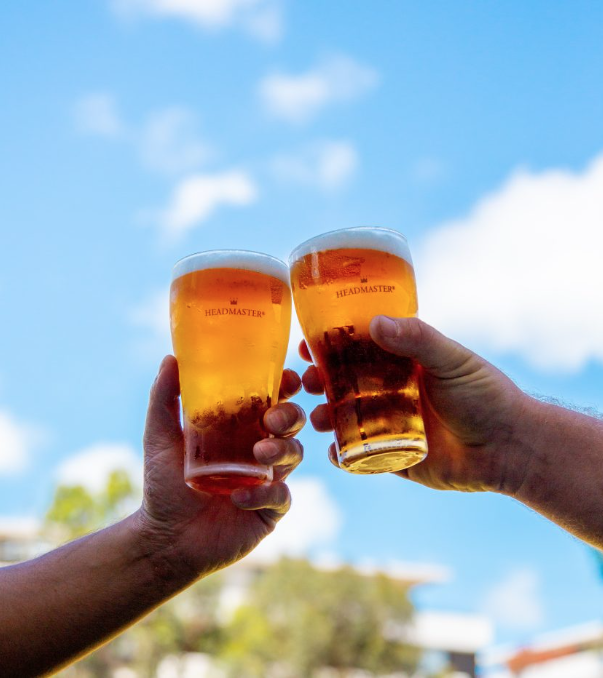 Two hands holding glasses of beer clinking together against a blue sky with clouds.