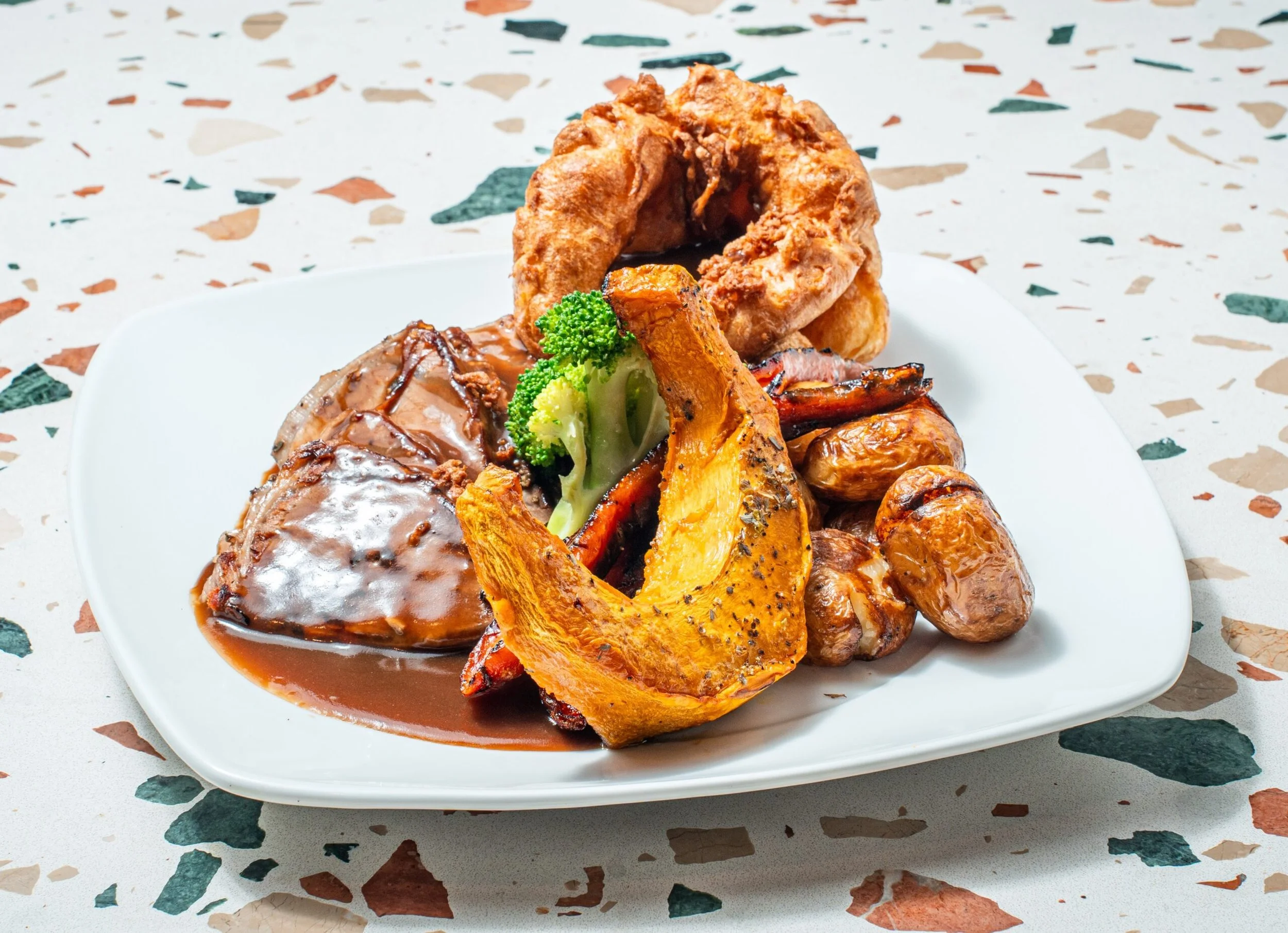 A white rectangular plate with a serving of meat with gravy, roasted vegetables including butternut squash, carrots, broccoli, and roasted potatoes.